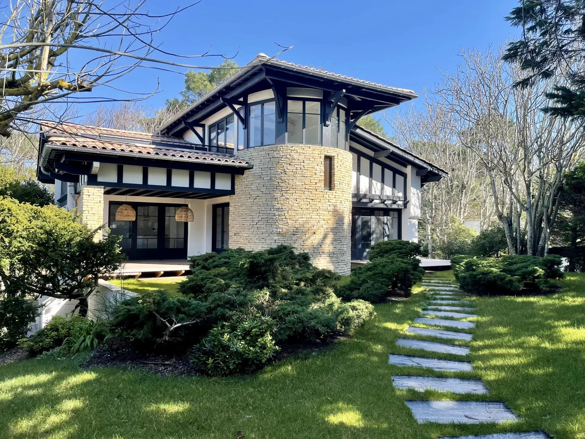 Maison moderne à deux étages avec un toit en pente, une tour ronde en pierre et un jardin bien entretenu avec un chemin en pierre.