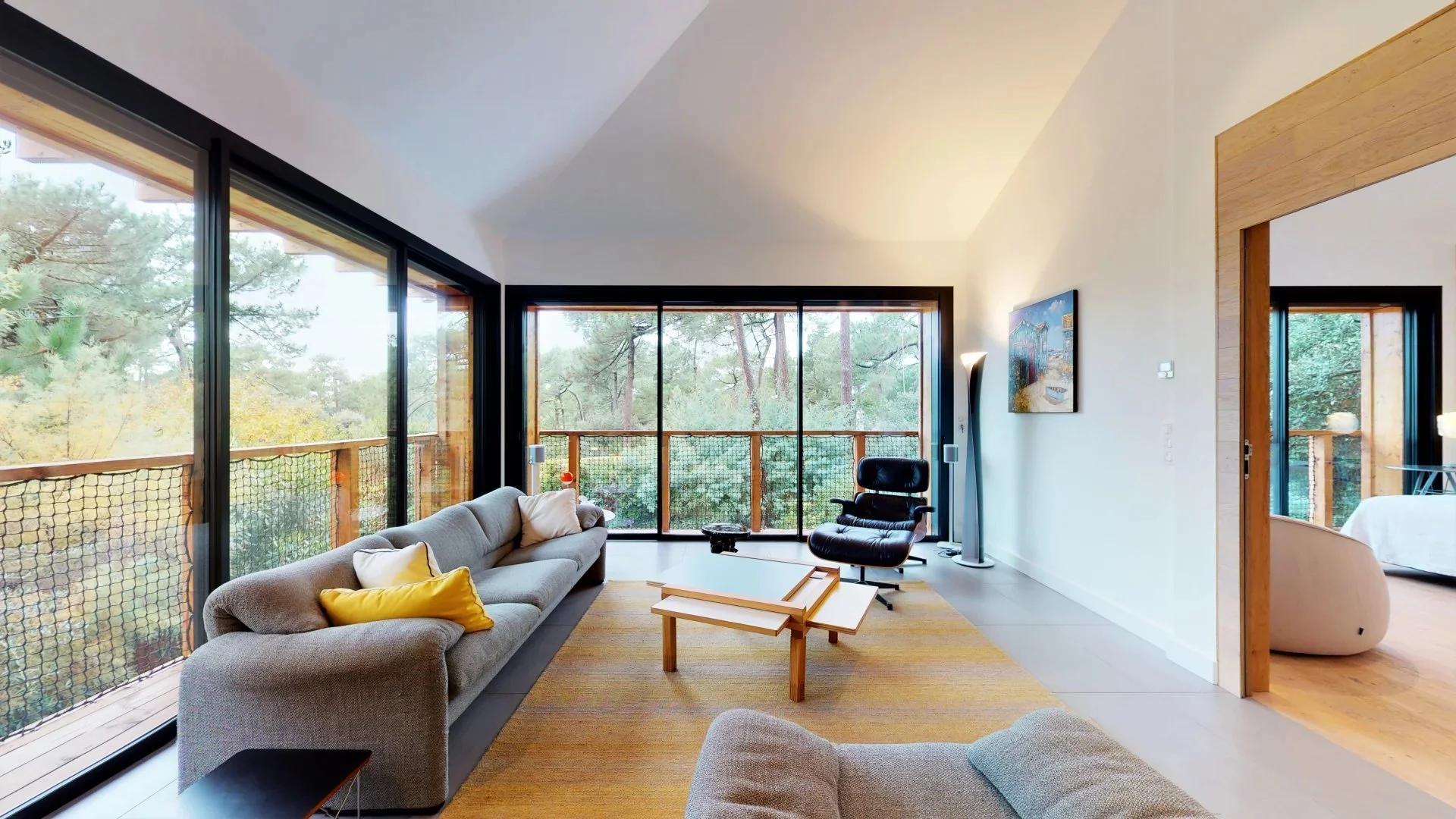 Intérieur d'un salon avec de grands carreaux en verre offrant une vue sur un bois, mobilier moderne comprenant un canapé gris, un fauteuil noir, des tables en bois clair, une peinture sur le mur, et une lampe sur pied, avec une porte menant à une autre pièce ayant également un grand accès à une terrasse ou un balcon en bois.