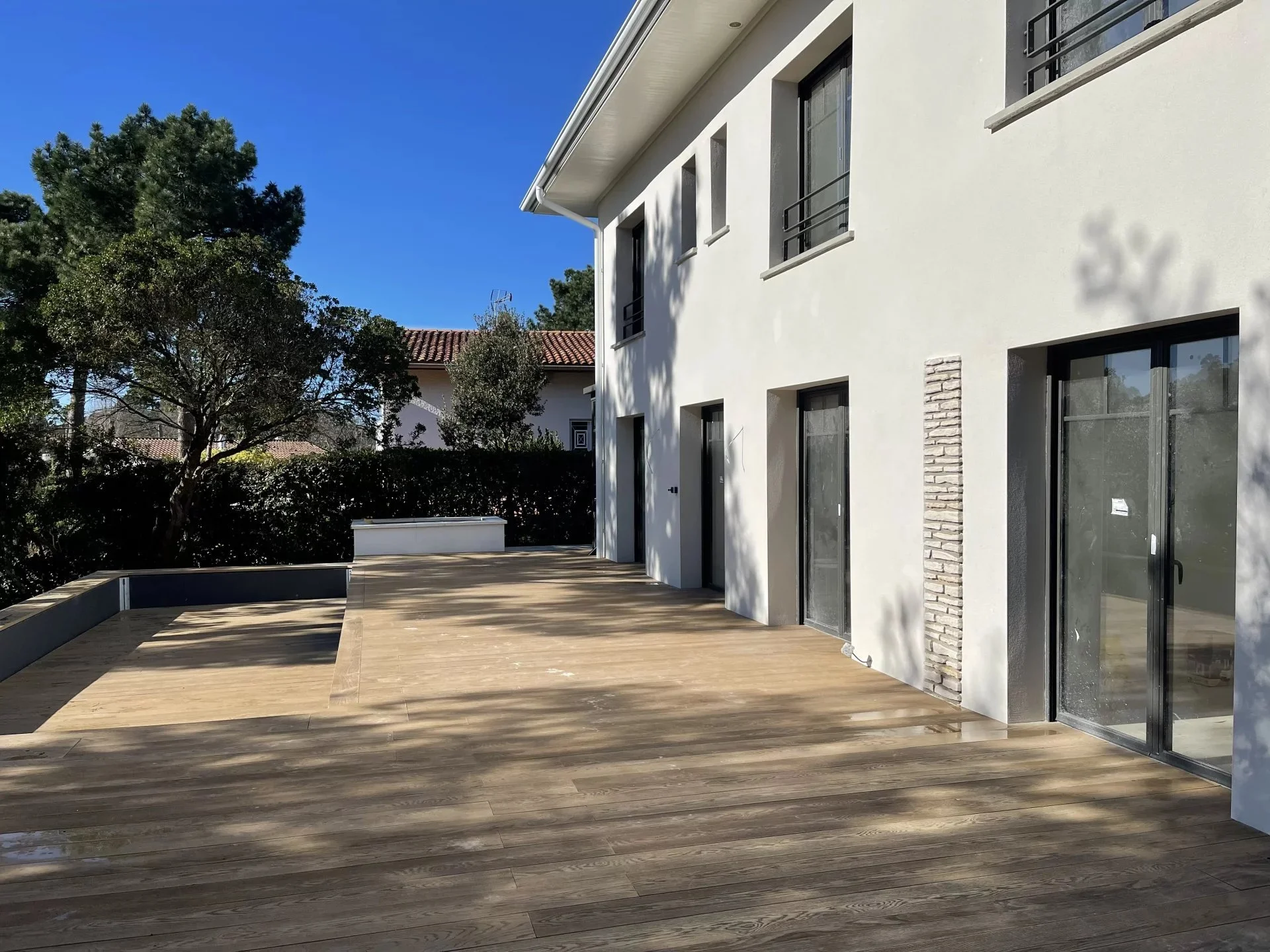 Vue d'une terrasse en bois adossée à un bâtiment moderne blanc avec plusieurs fenêtres et portes vitrées, entourée de végétation et sous un ciel bleu clair.