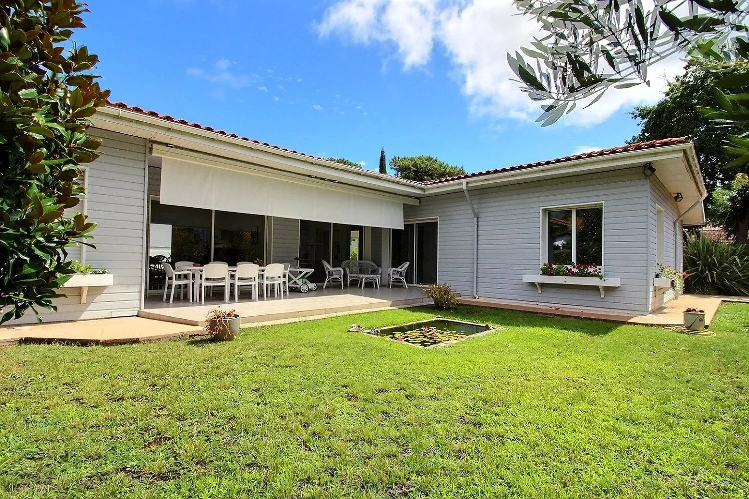 Maison de plain-pied avec terrasse, jardin, piscine gonflable et mobilier de jardin, sous un ciel bleu avec quelques nuages