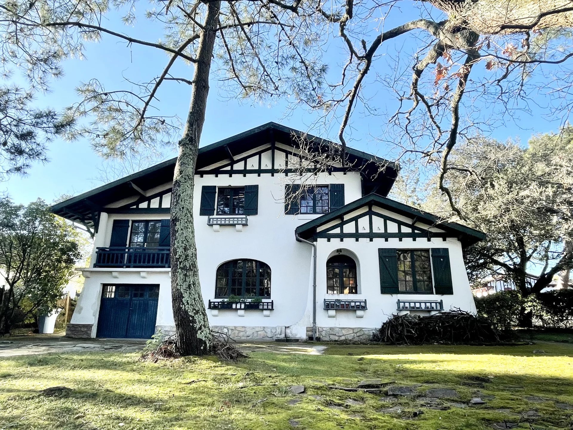 Maison blanche avec volets noirs, entourée d'arbres au printemps, sous un ciel bleu.