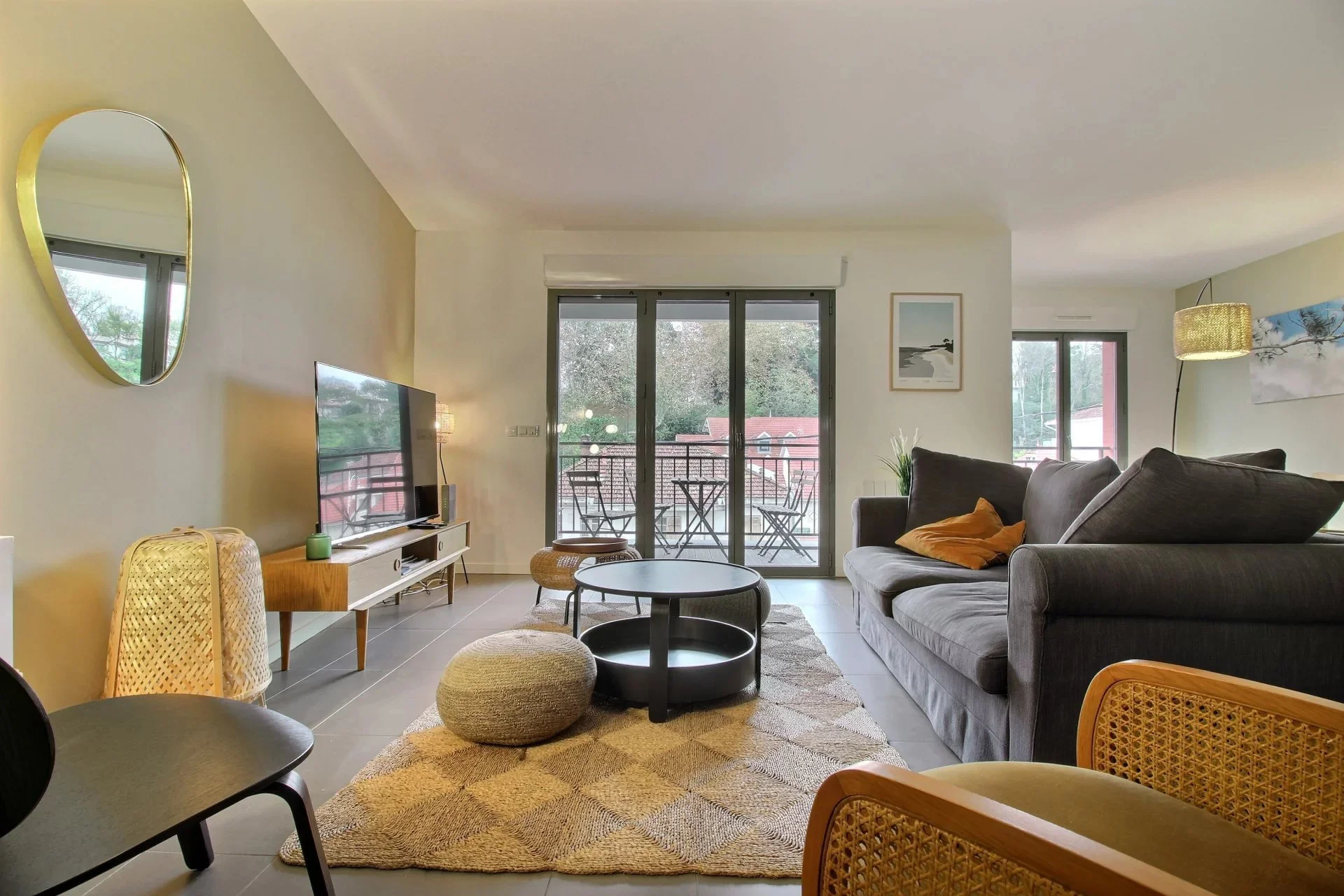 Salon avec canapé gris, télévision sur meuble blanc, tapis beige, grande baie vitrée avec balcon et mobilier extérieur, décor avec tableaux et lampes.