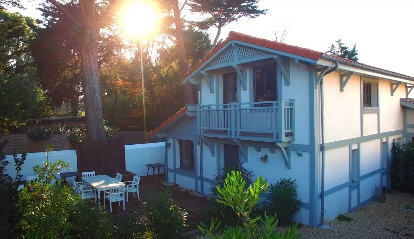 Maison de style cottage à deux étages avec balcon, entourée d'arbres et de plantes, avec une table et des chaises dans la cour, sous un soleil brillant.