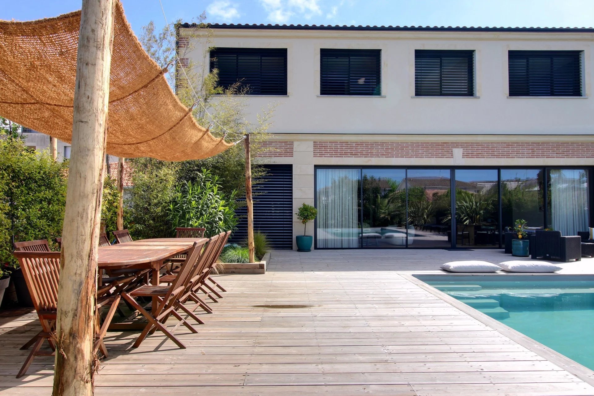 Une terrasse en bois avec une piscine, une table en bois et des chaises, des plantes en pot et un bâtiment moderne avec de grandes fenêtres en arrière-plan.