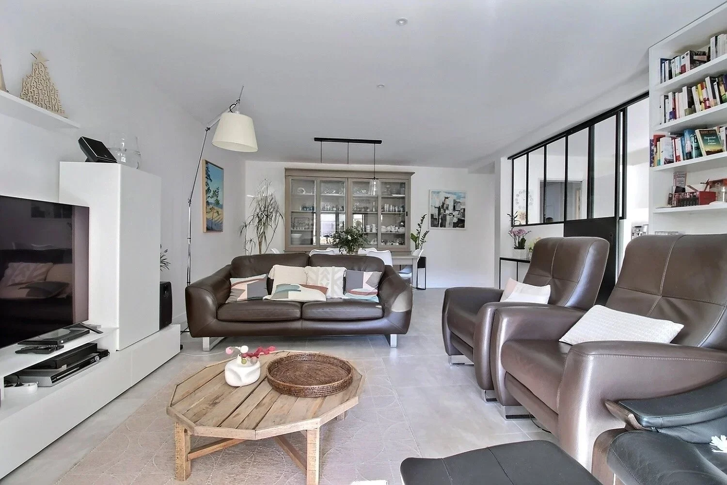 Salon moderne avec canapé en cuir marron, fauteuils en cuir gris, table basse en bois, étagères blanches remplies de livres, une grande armoire en verre au fond, et une cloison en métal noir séparant l'espace.