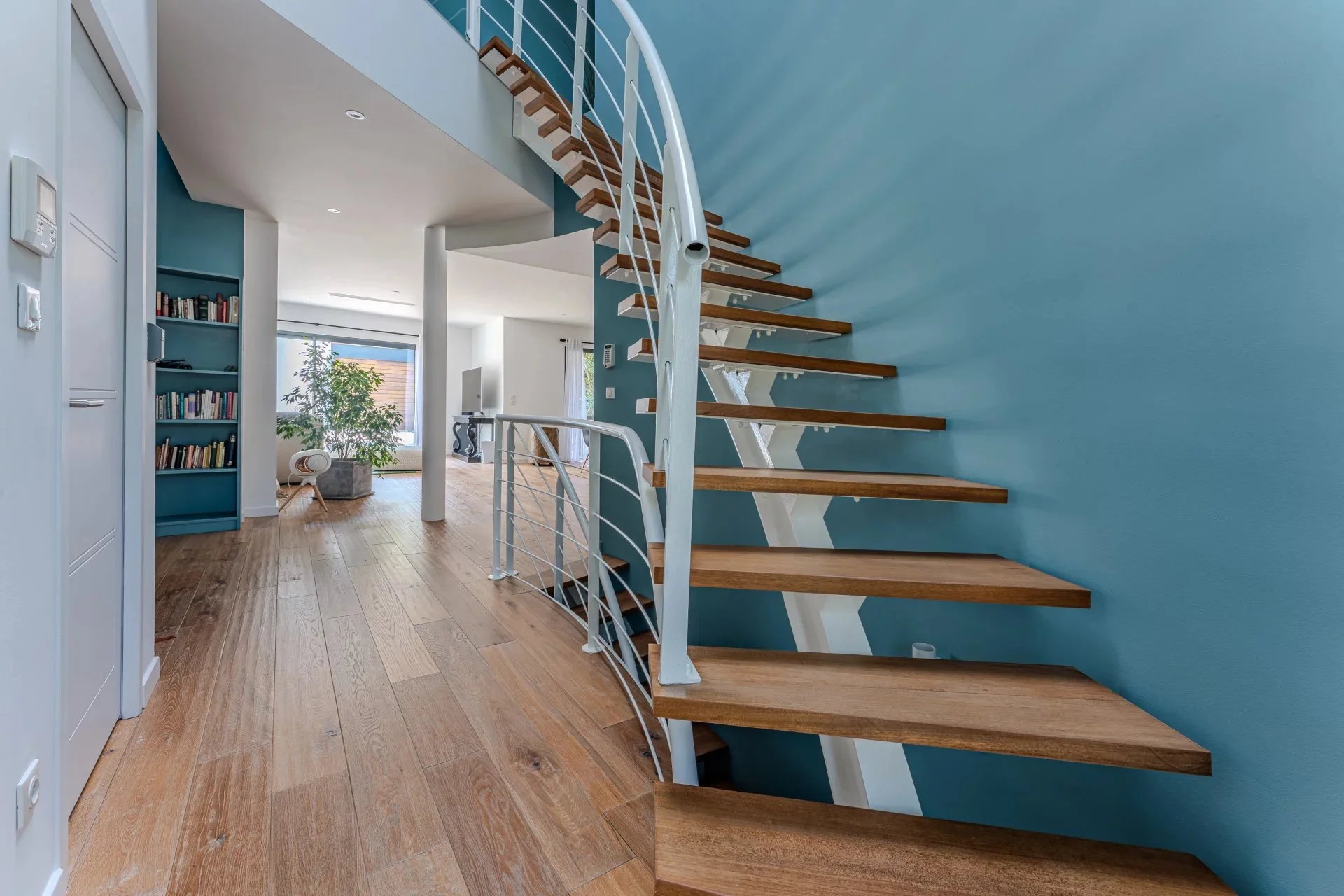 Escalier intérieur en bois avec main courante en métal, mur bleu derrière, salon lumineux avec bibliothèque et plantes.