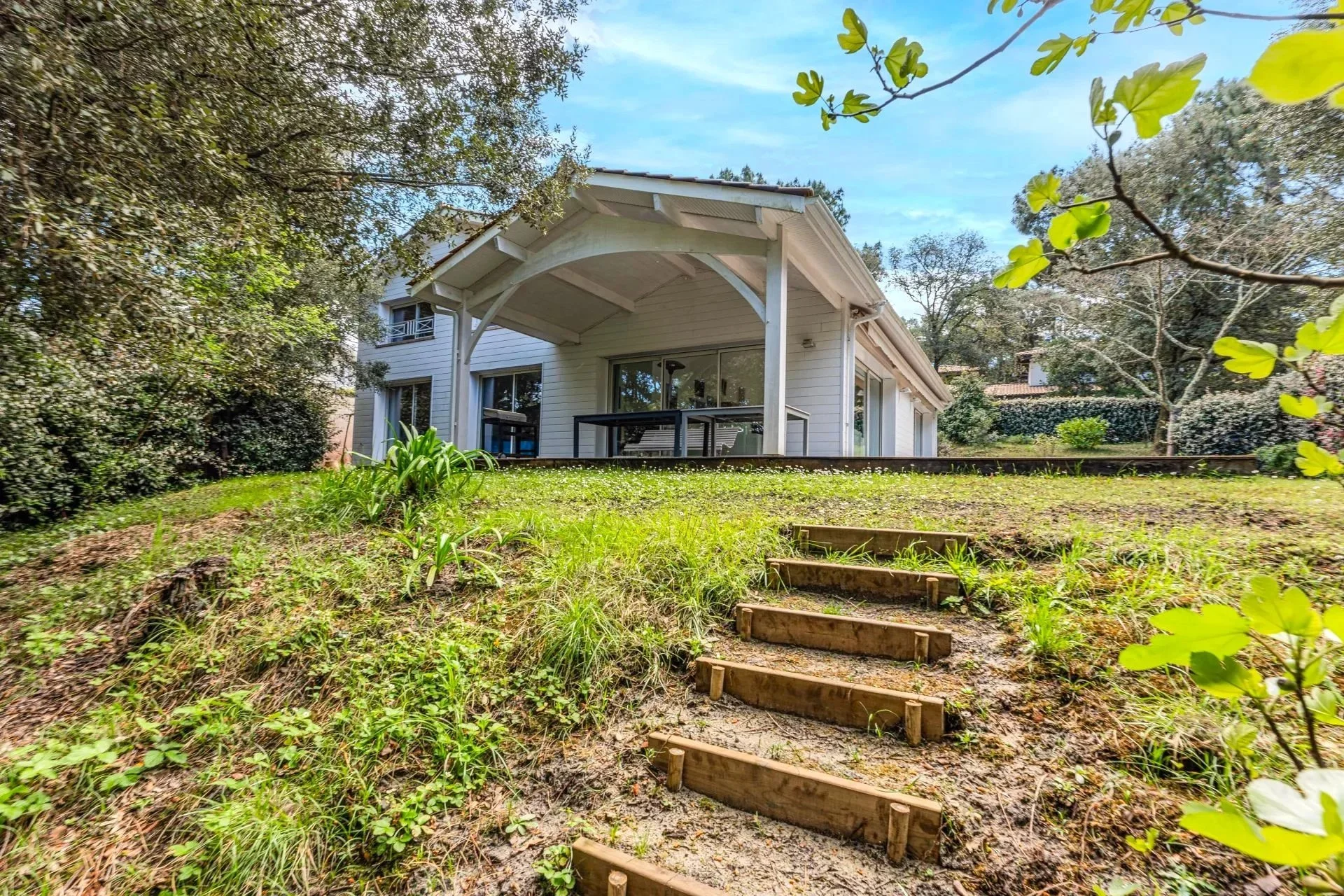 Maison blanche avec une terrasse sous un auvent, vue depuis un jardin avec des escaliers en bois menant à la maison, entouré d'arbres et de végétation.