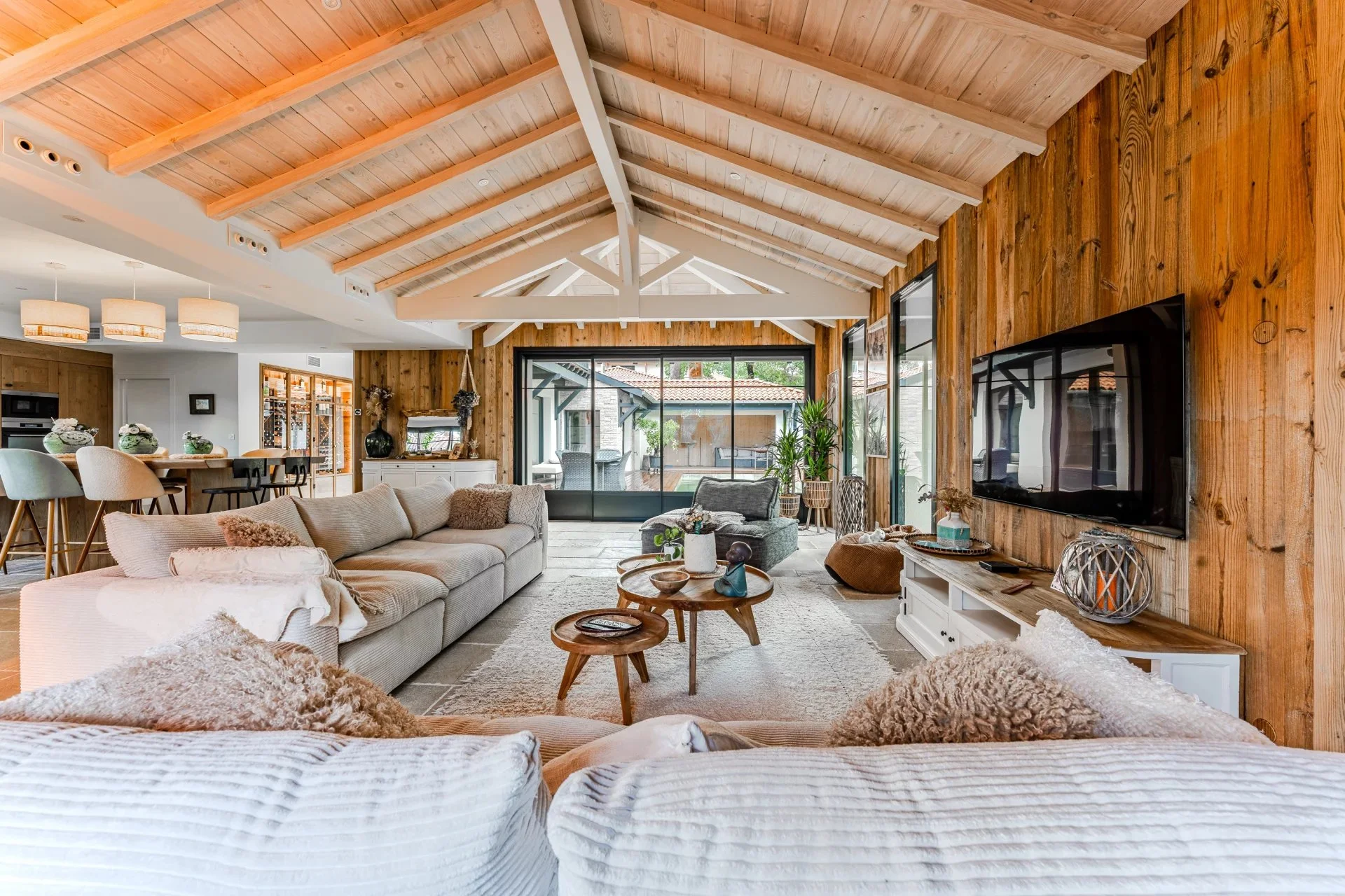 Intérieur d’un salon avec un plafond en bois, un mur en bois, un canapé beige, une table basse en bois, une télévision sur le mur et des plantes d’intérieur. Une grande porte-fenêtre en verre donne sur une terrasse extérieure.