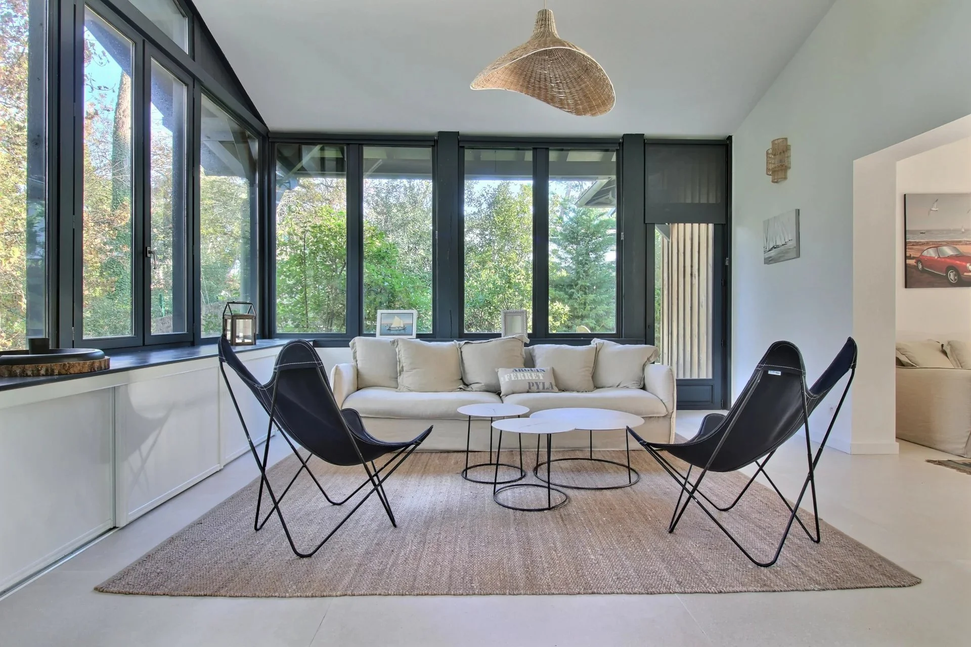 Salon intérieur avec grand fenêtres, canapé blanc, chaises noires, tapis, décorations murales et vue sur un espace extérieur avec des arbres.