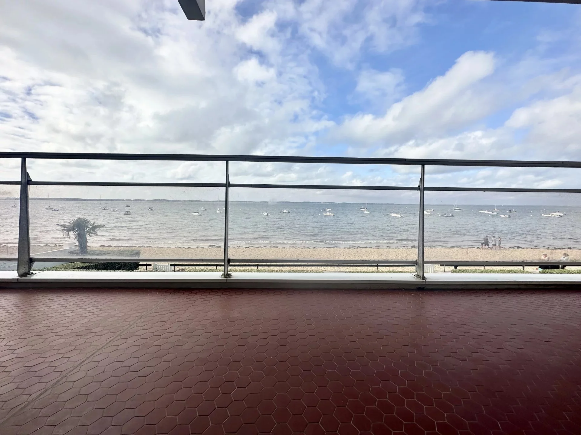 Vue depuis un balcon en terrasse avec un sol en hexagones rouges, regardant une plage, des bateaux et deux personnes avec un chien sur la plage, sous un ciel partiellement nuageux.