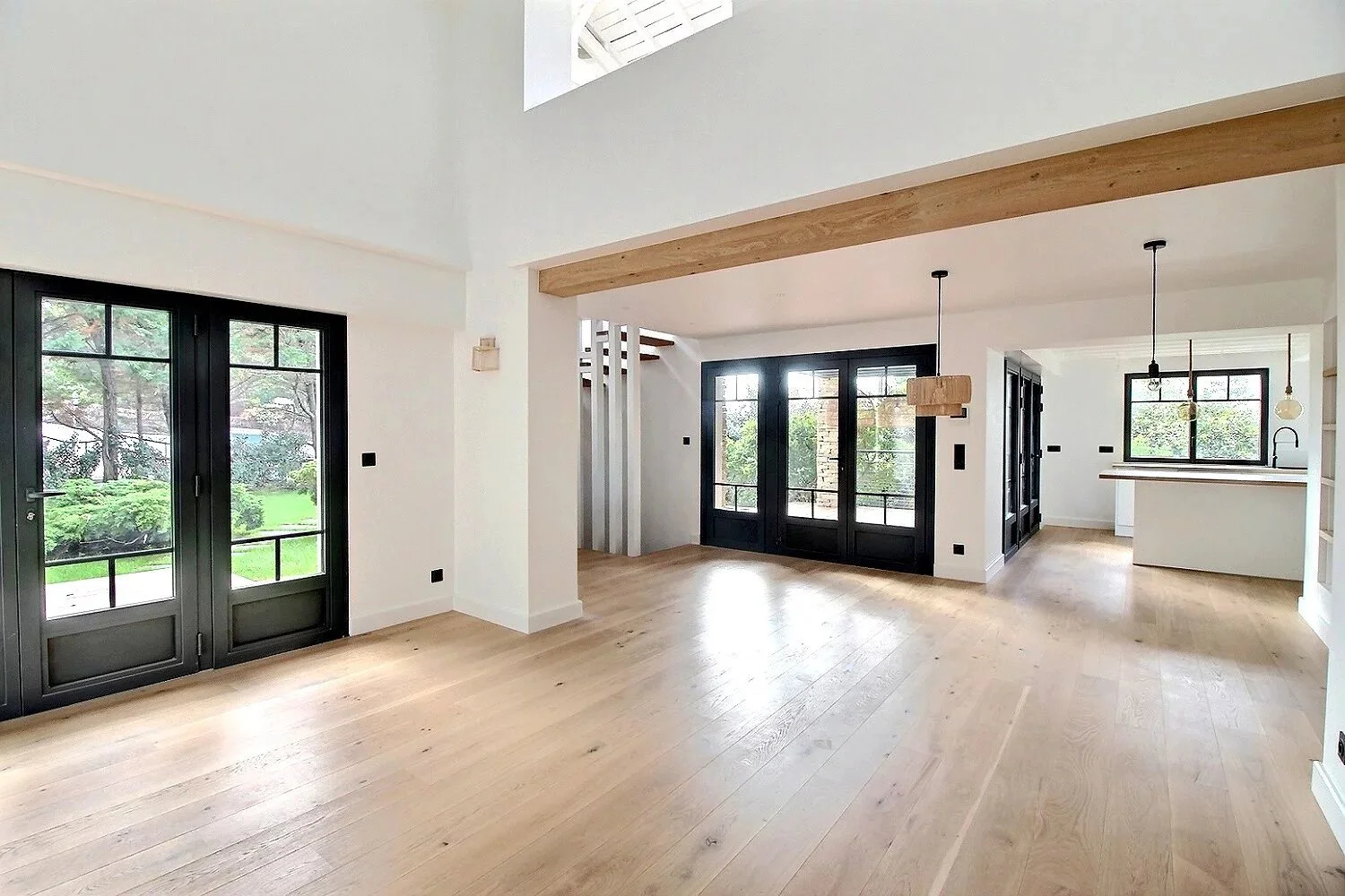 Intérieur d'une pièce lumineuse avec sol en bois clair, murs blancs, grandes fenêtres avec cadre noir, et porte-fenêtre donnant sur un jardin vert. Espace ouvert avec poutres en bois et luminaires suspendus, vue vers la cuisine.