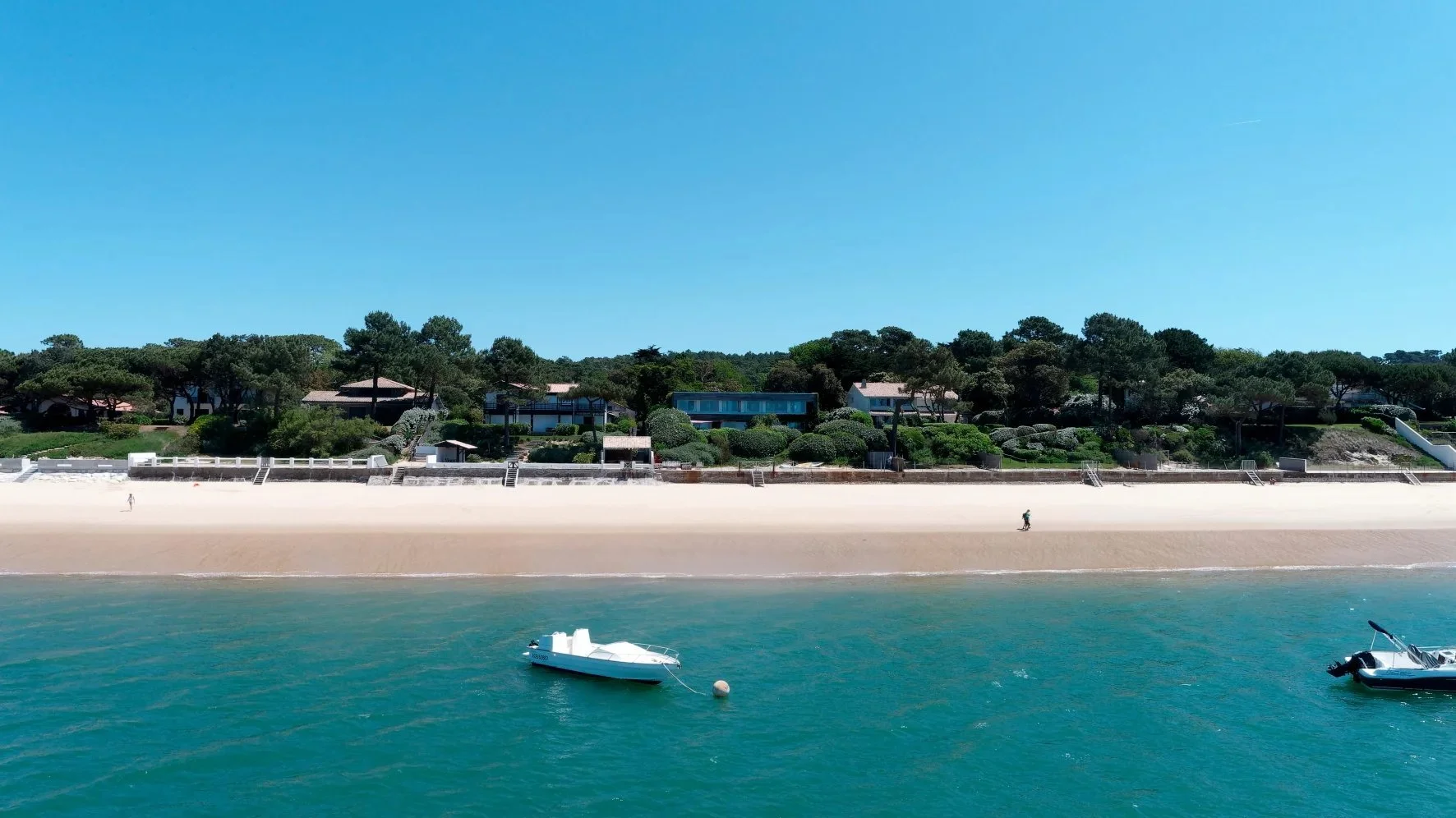 Plage de sable fin avec deux bateaux à flottant, bordée de maisons et de végétation luxuriante, sous un ciel bleu clair.