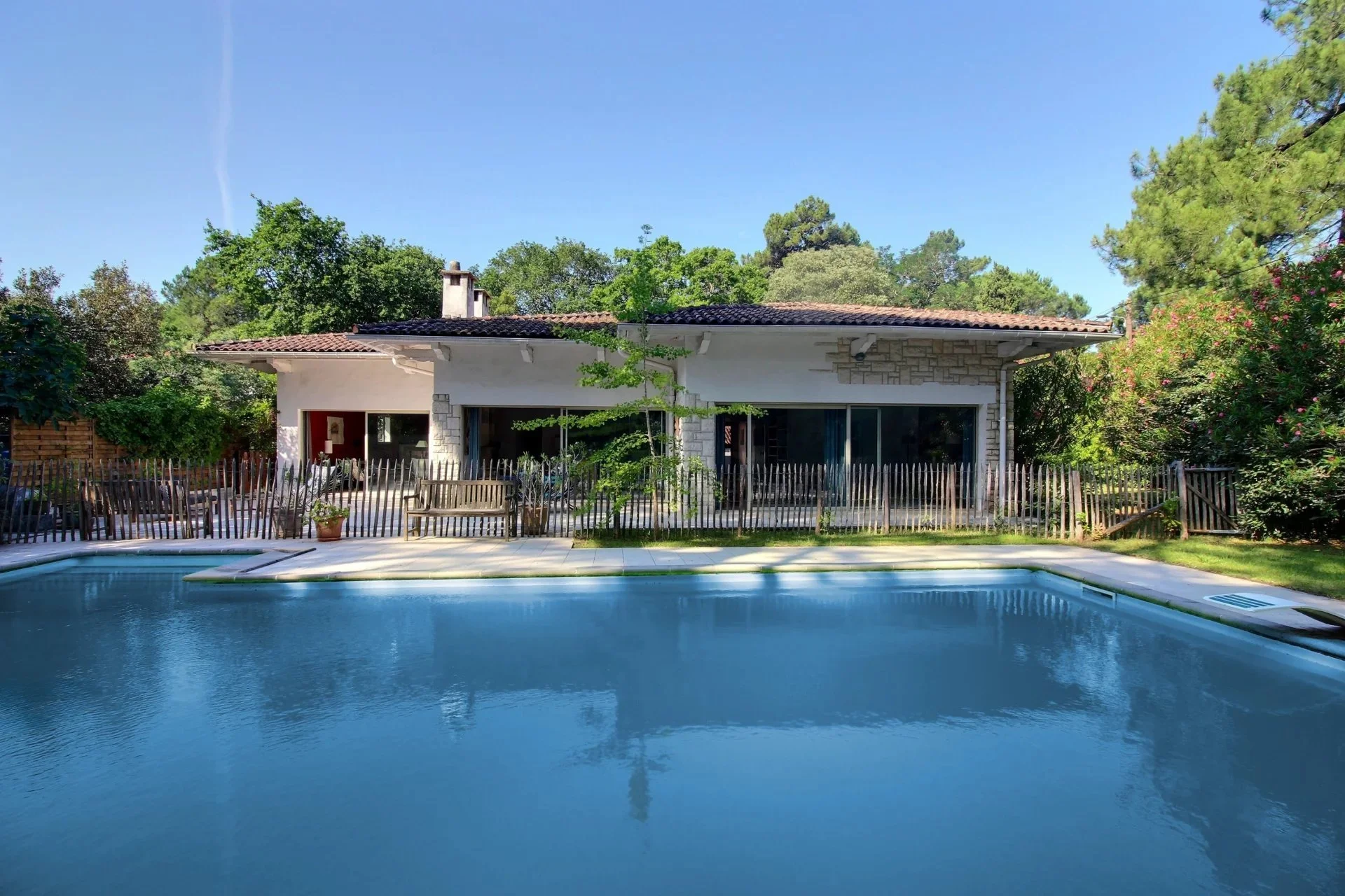 Une maison de plain-pied avec une piscine devant, entourée de végétation et de arbres, sous un ciel clair.
