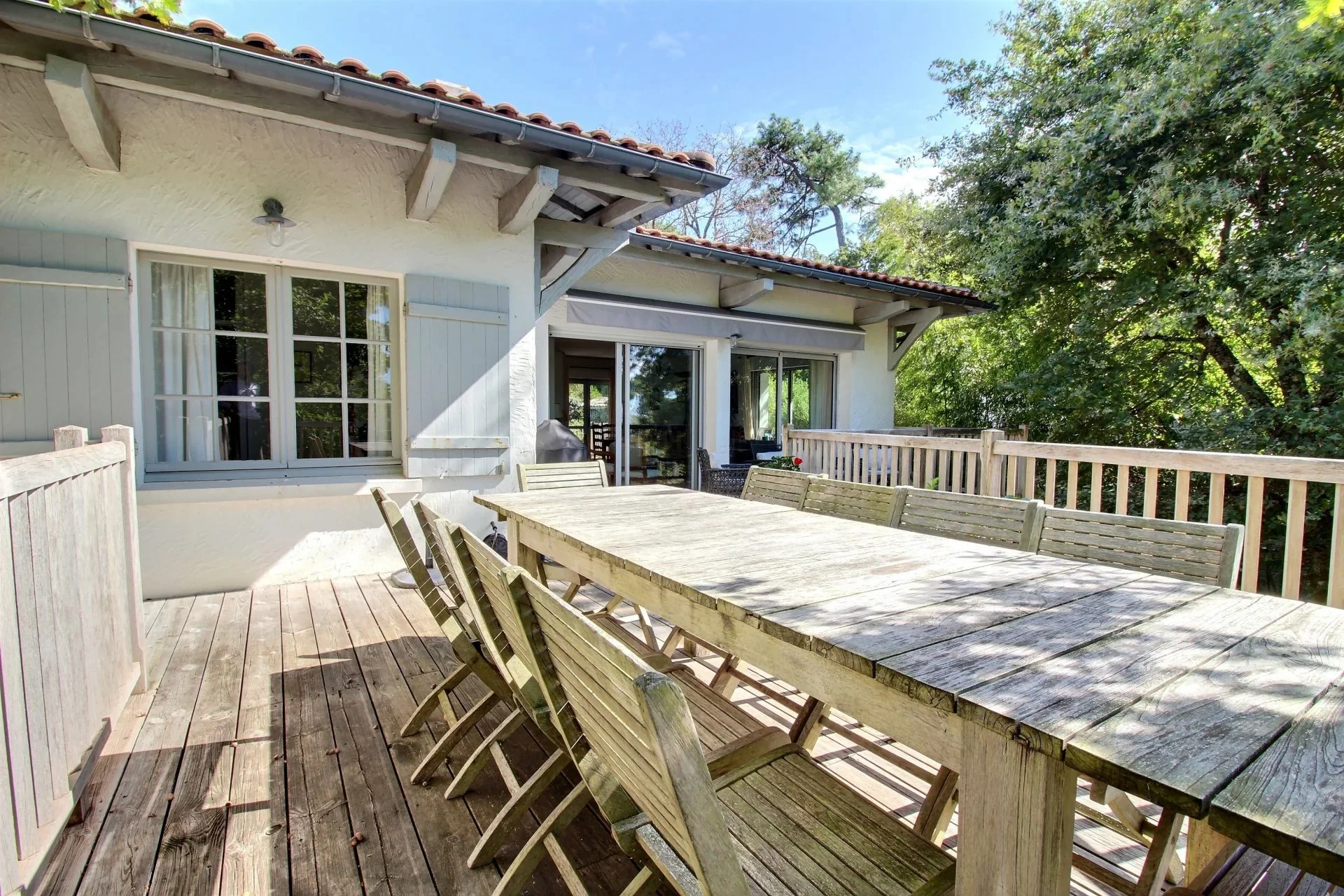 Terrasse en bois avec une grande table et des chaises en bois, entourée de panneaux en bois, avec une maison blanche en arrière-plan, entourée d'arbres verts et sous un ciel bleu.