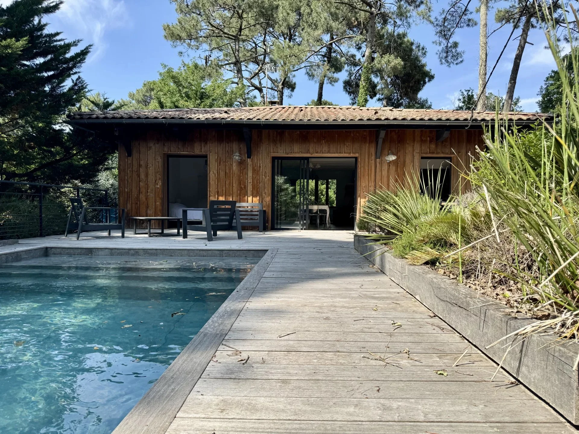 Maison en bois avec une terrasse en bois, à côté d'une piscine, entourée d'arbres et de plantes, sous un ciel bleu.
