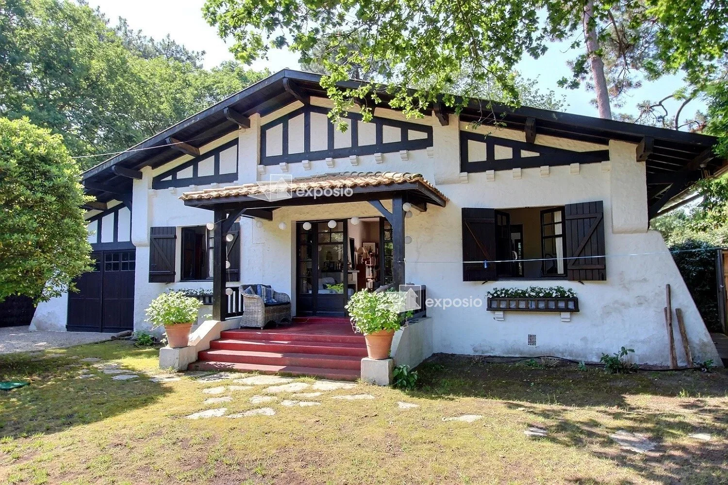 Maison avec façade blanche, volets noirs, et terrasse avec escaliers rouges, entourée d'arbres et de végétation.