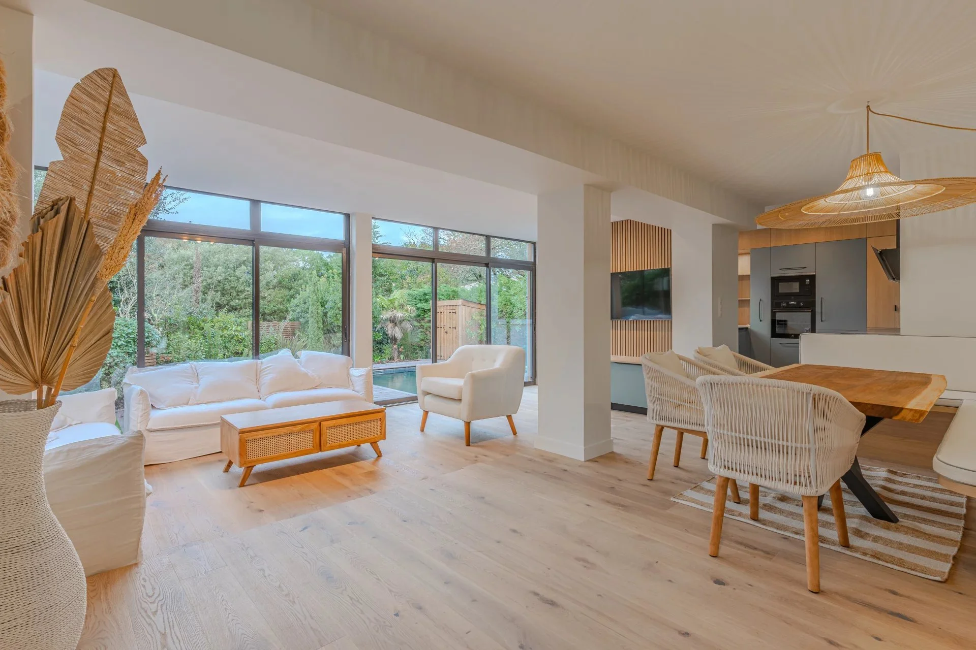 Salon lumineux avec canapés blancs, fenêtres en verre, vue sur un jardin, mobilier en bois clair et décoration naturelle.