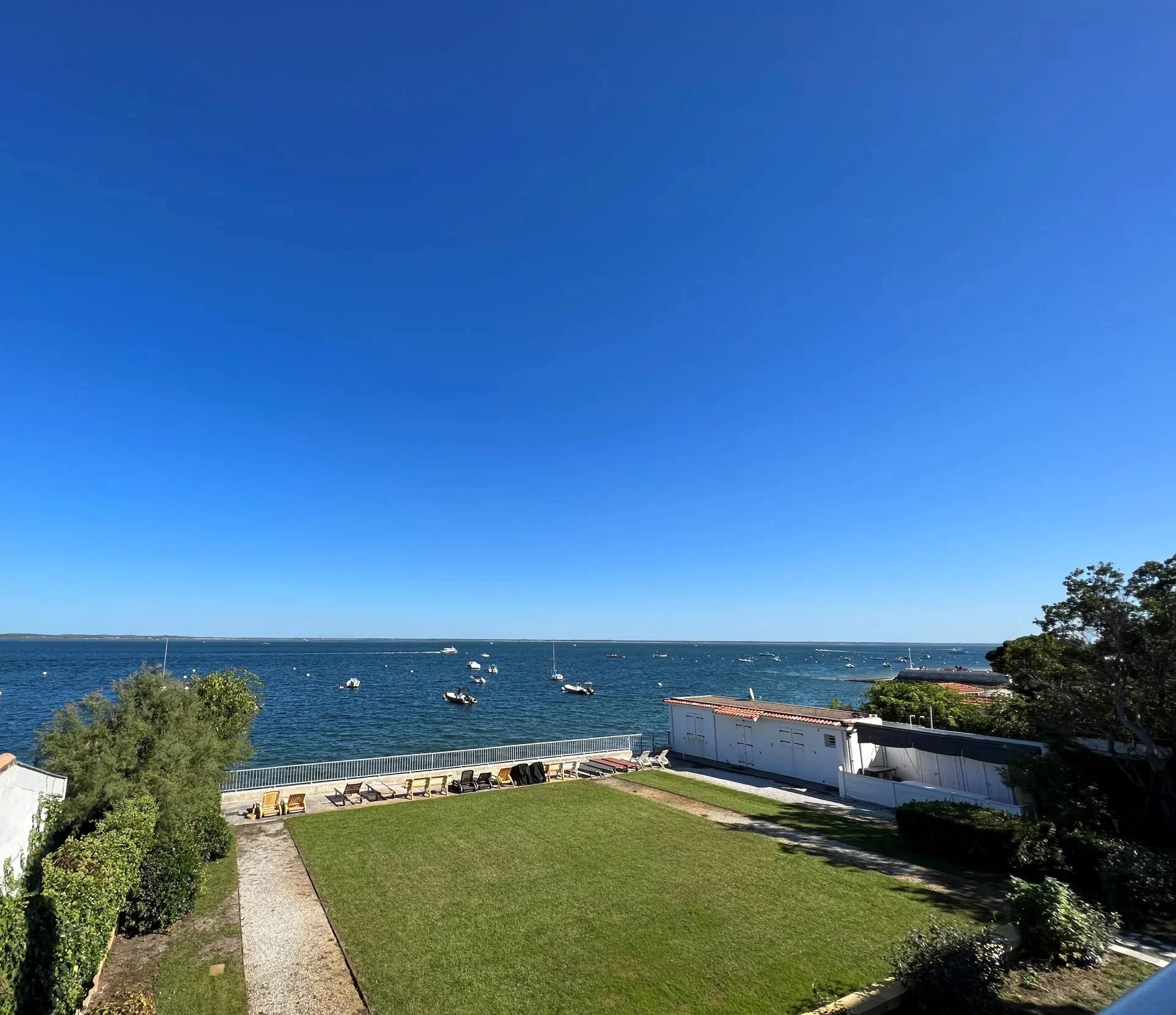 Vue sur un ciel bleu clair au-dessus d'une mer avec plusieurs bateaux, vue depuis une propriété avec un jardin vert et quelques petits bâtiments blancs.