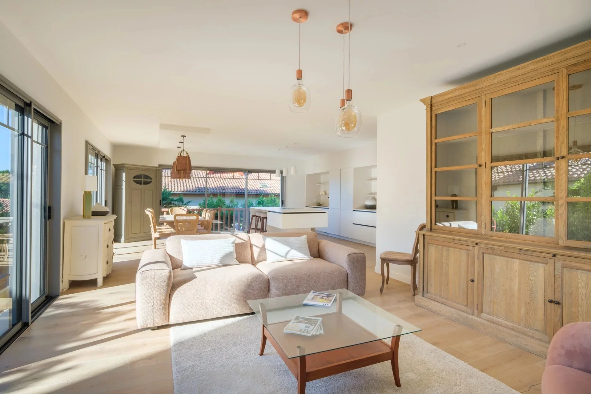 Intérieur d'un salon lumineux avec canapé beige, table en verre, bibliothèque en bois, et vue sur un balcon avec des plantes et un toit en tuiles.