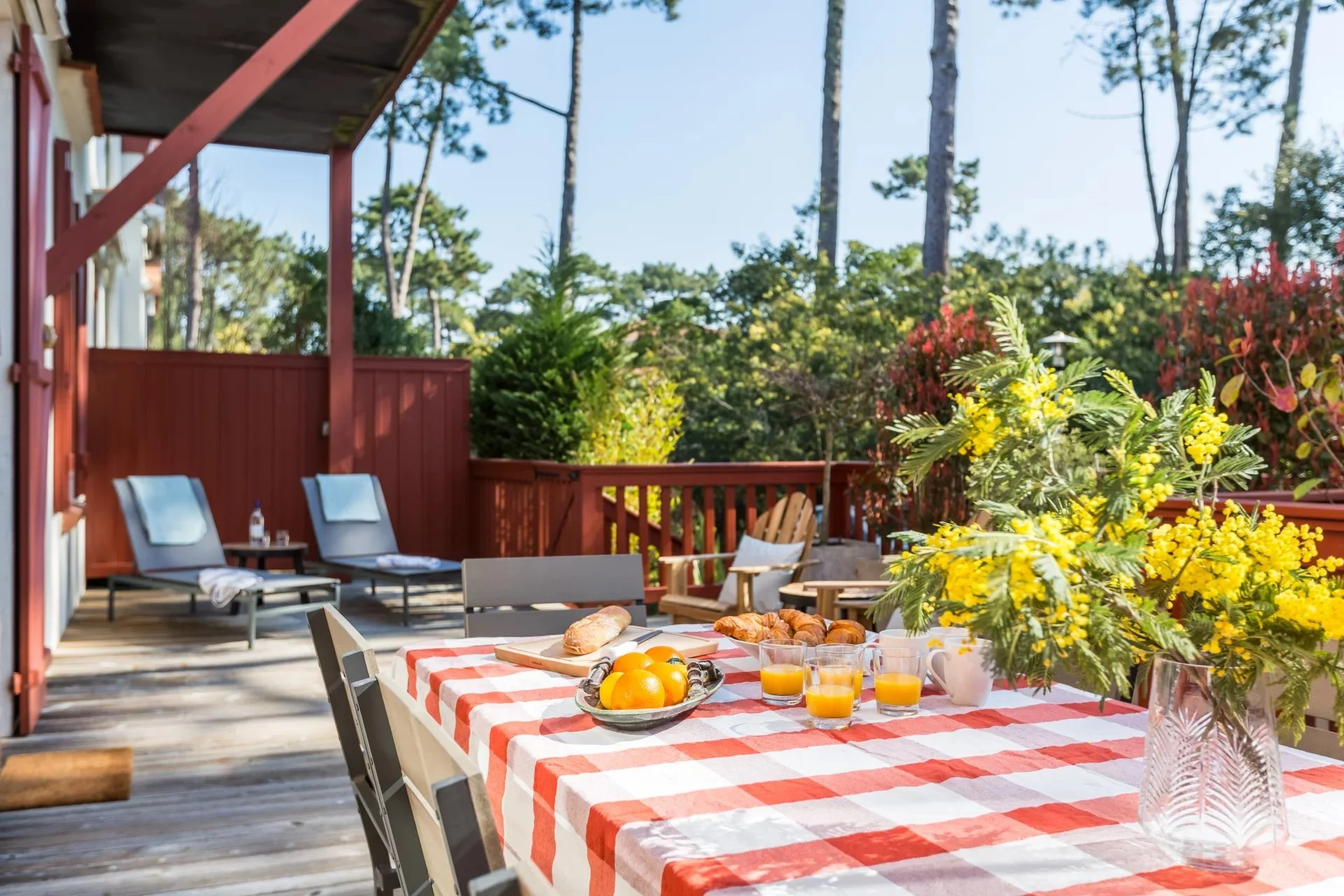 Repas de petit-déjeuner en plein air sur une terrasse en bois avec vue sur la forêt, comprenant un tableau à carreaux rouges et blancs avec des croissants, des oranges, des verres de jus d'orange, une tasse, une carafe et un bouquet de fleurs jaunes.