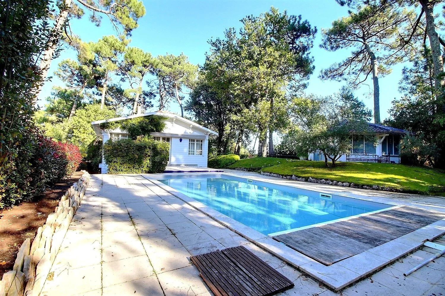 Piscine extérieure dans un jardin boisé avec deux petites maisons en arrière-plan, entourée d'arbres et de pelouse verte, par une journée ensoleillée.