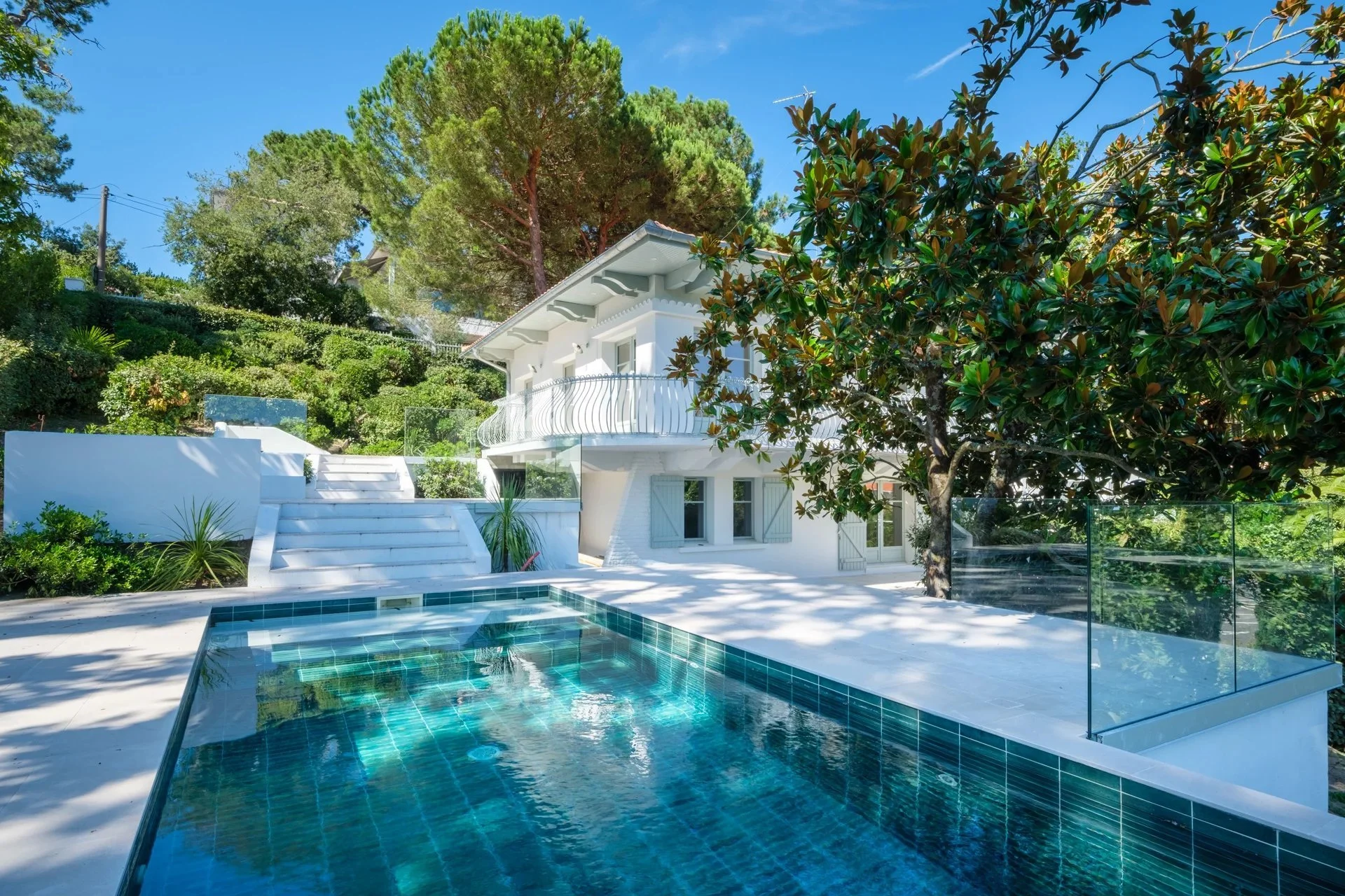 Maison blanche avec piscine à l'extérieur, entourée d'arbres et de végétation, sous un ciel bleu clair.