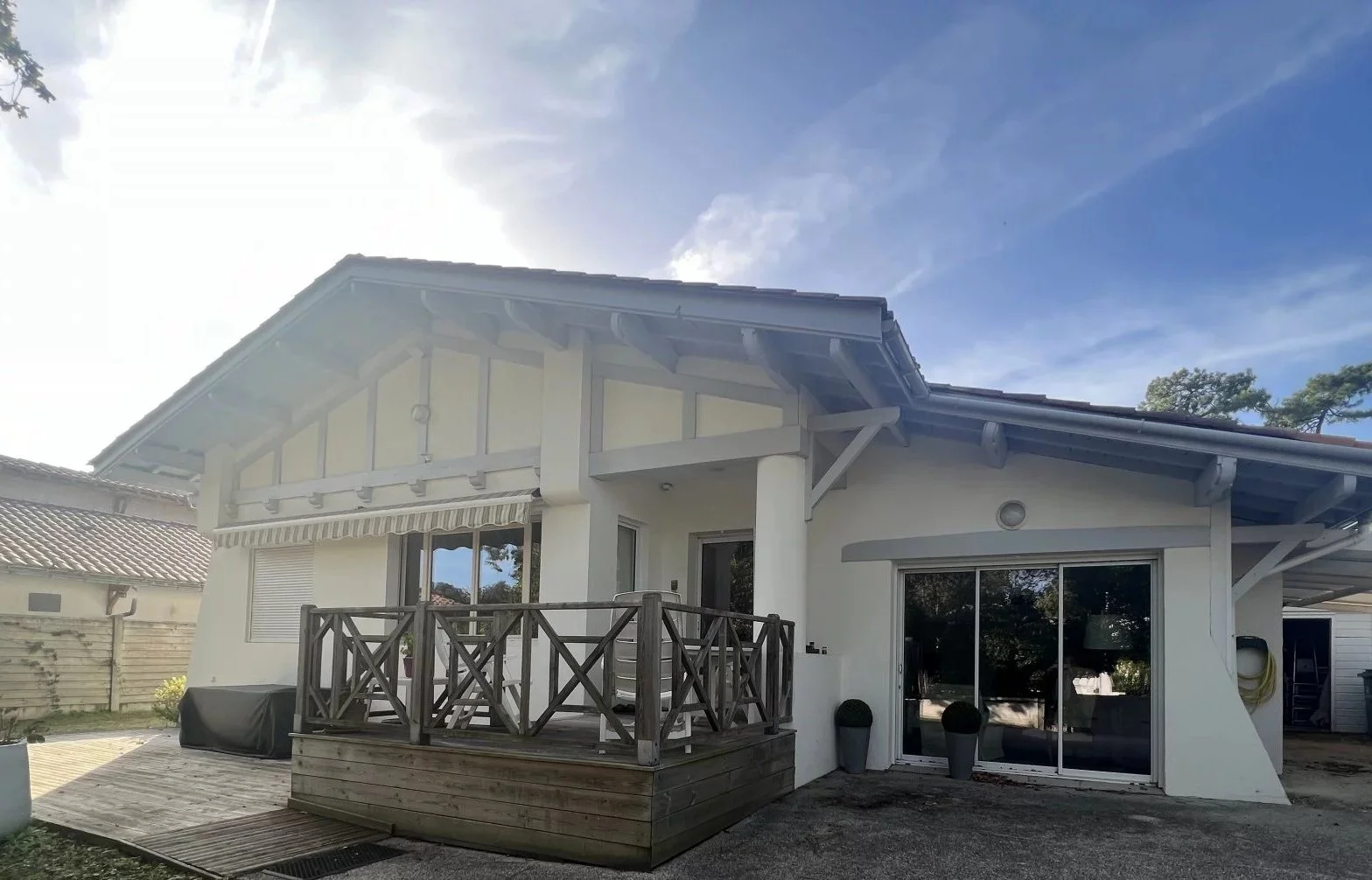 Maison moderne blanche avec un balcon en bois et des fenêtres vitrées, sous un ciel bleu avec quelques nuages.