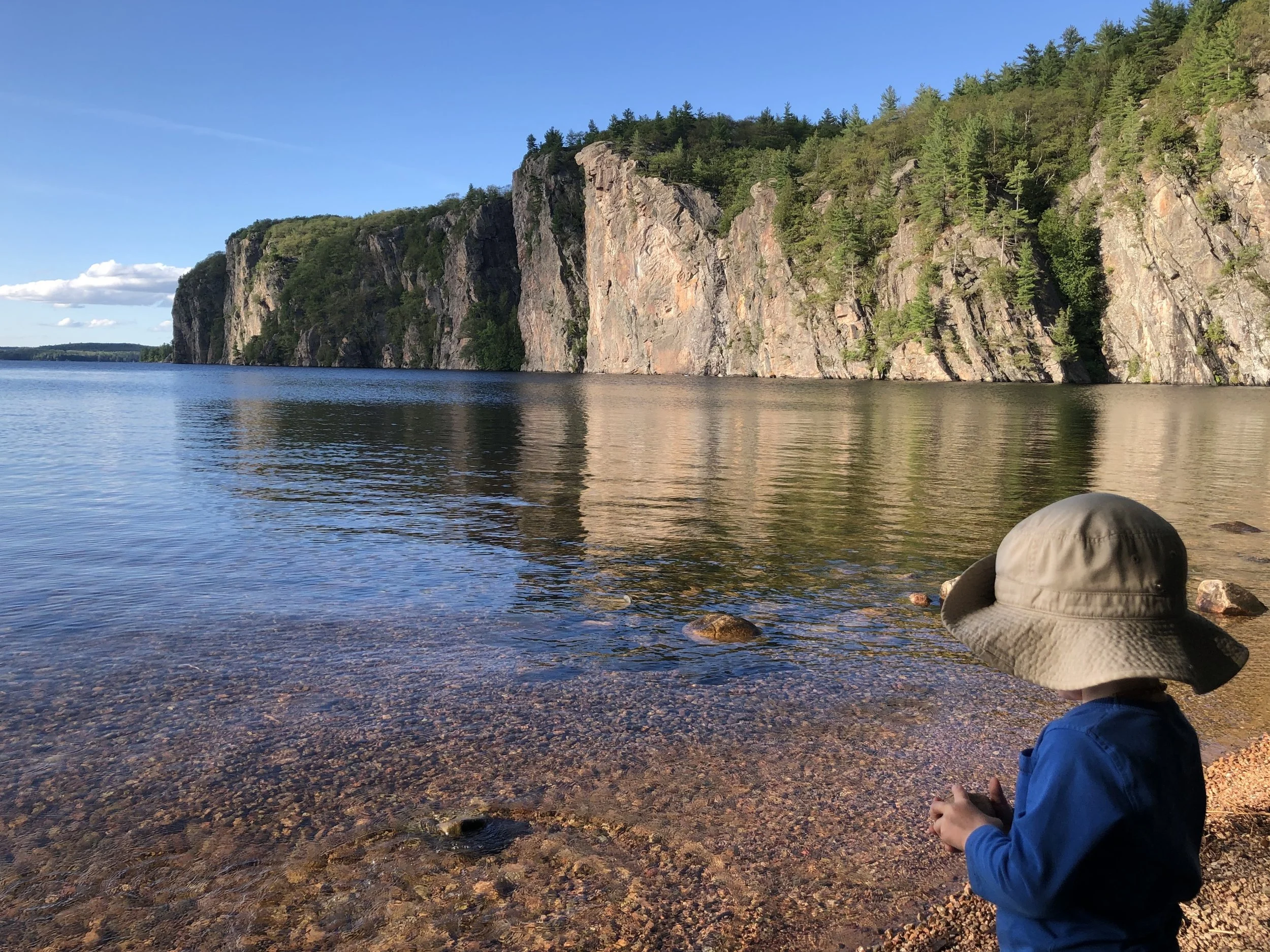 A child with a wide-brimmed hat and blue jacket standing at the rocky shore of a lake, with cliffs and green trees in the background on a clear day.