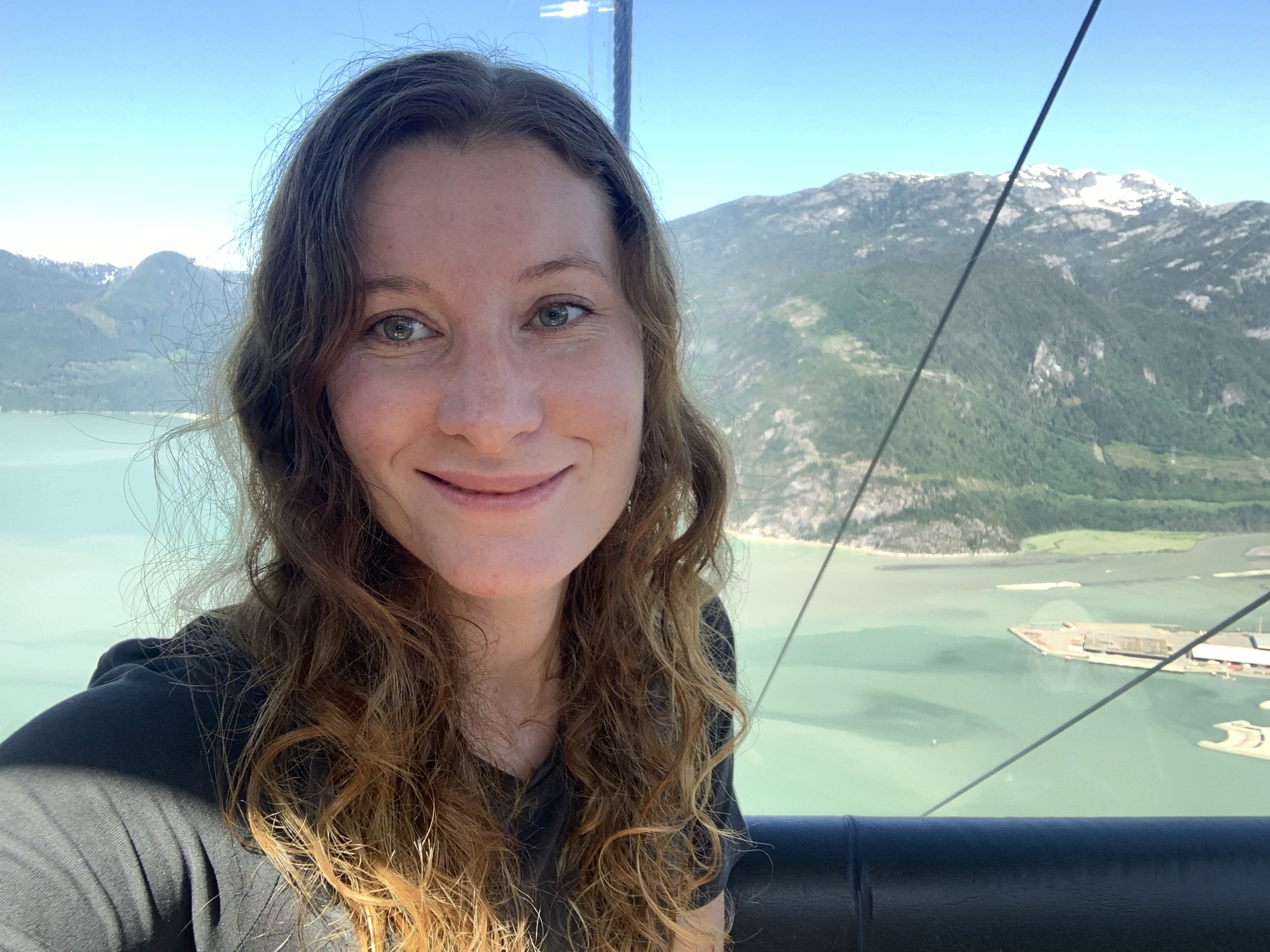 A woman with long curly hair smiling in front of a scenic mountain and water landscape from a gondola or cable car.