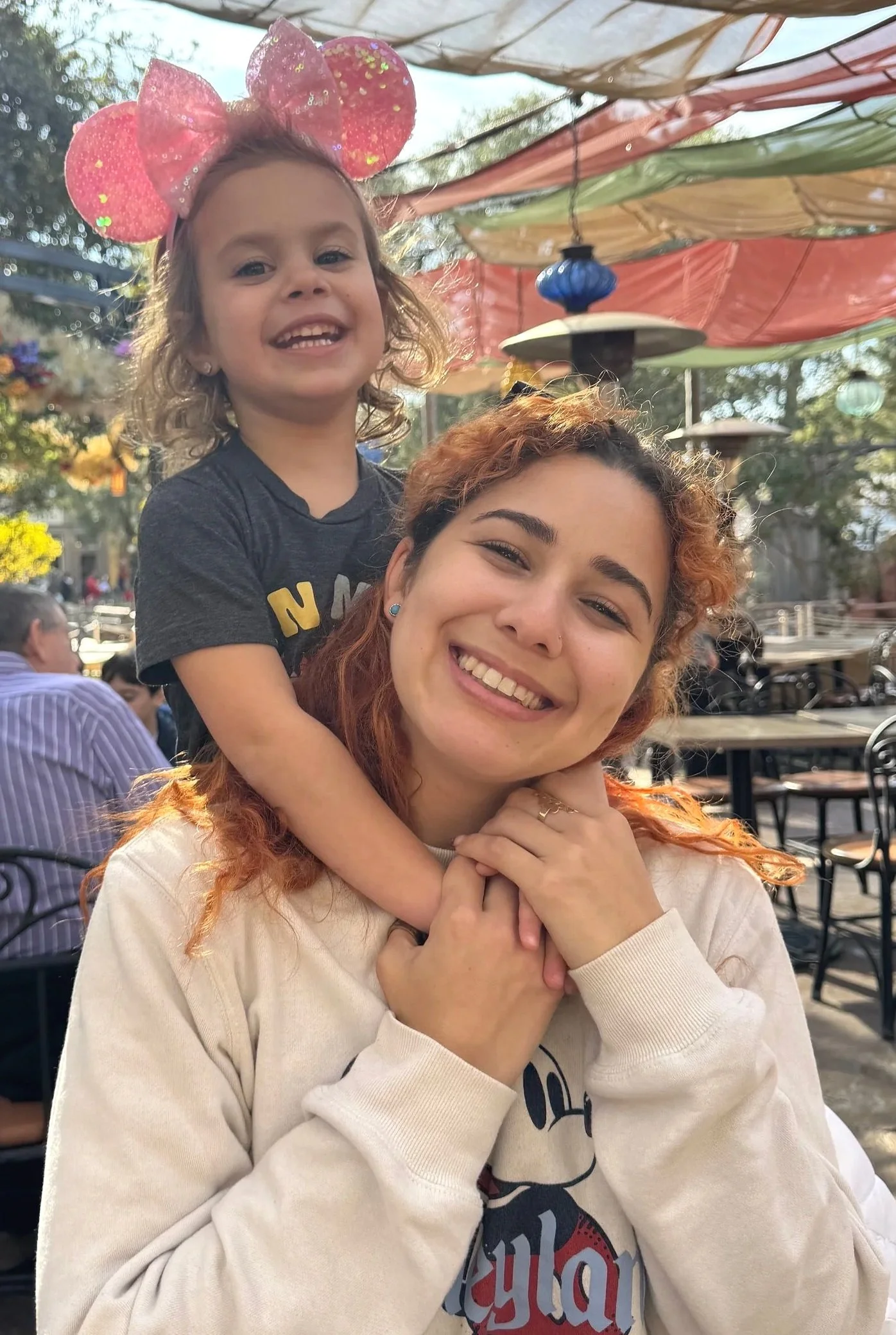 A woman with curly reddish hair smiling and holding a young girl with curly blonde hair on her shoulders. The girl is wearing a pink bow headband with sparkles and a dark gray shirt. The scene is outdoors at a patio with colorful umbrellas overhead, and there are other people in the background.