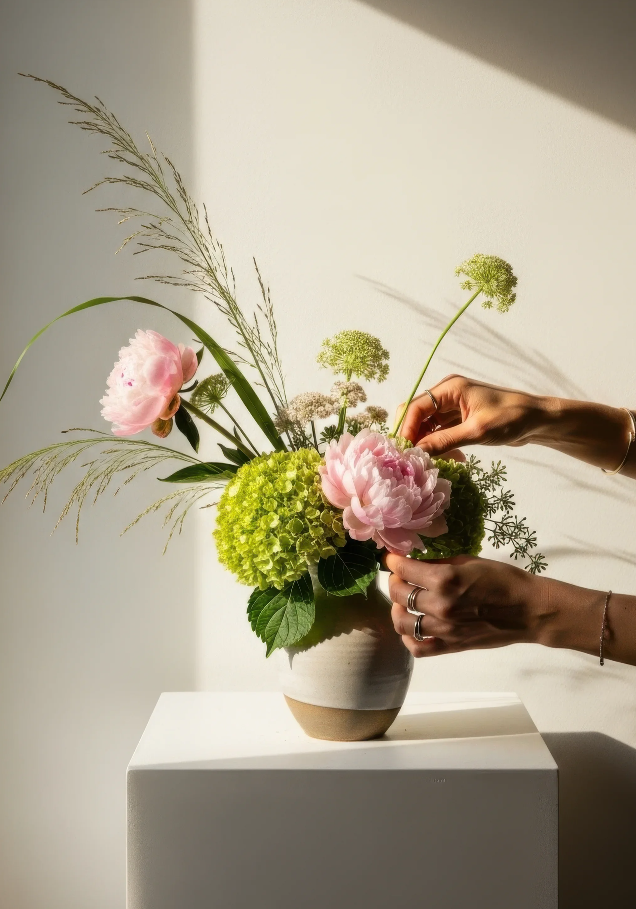 Person holding a floral arrangement with pink peonies, green hydrangeas, and assorted greenery, in a two-tone ceramic vase, against a plain wall with sunlight casting shadows.