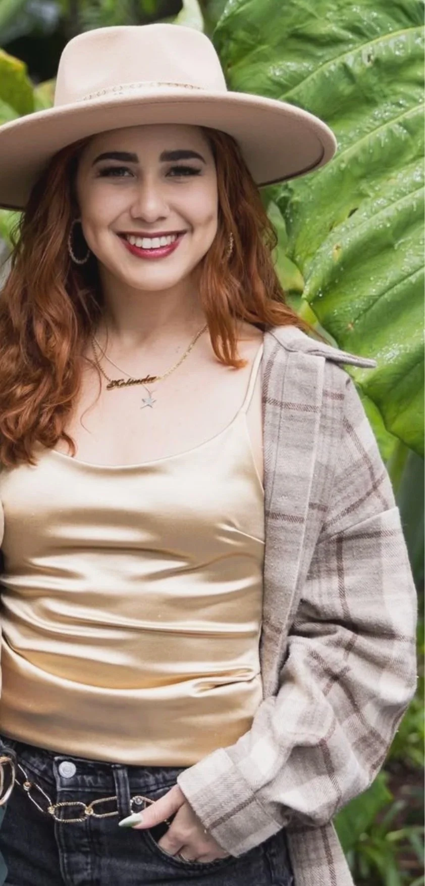A young woman smiling outdoors, wearing a beige hat, a shiny beige top, a plaid blazer, and jewelry, with large green leaves in the background.