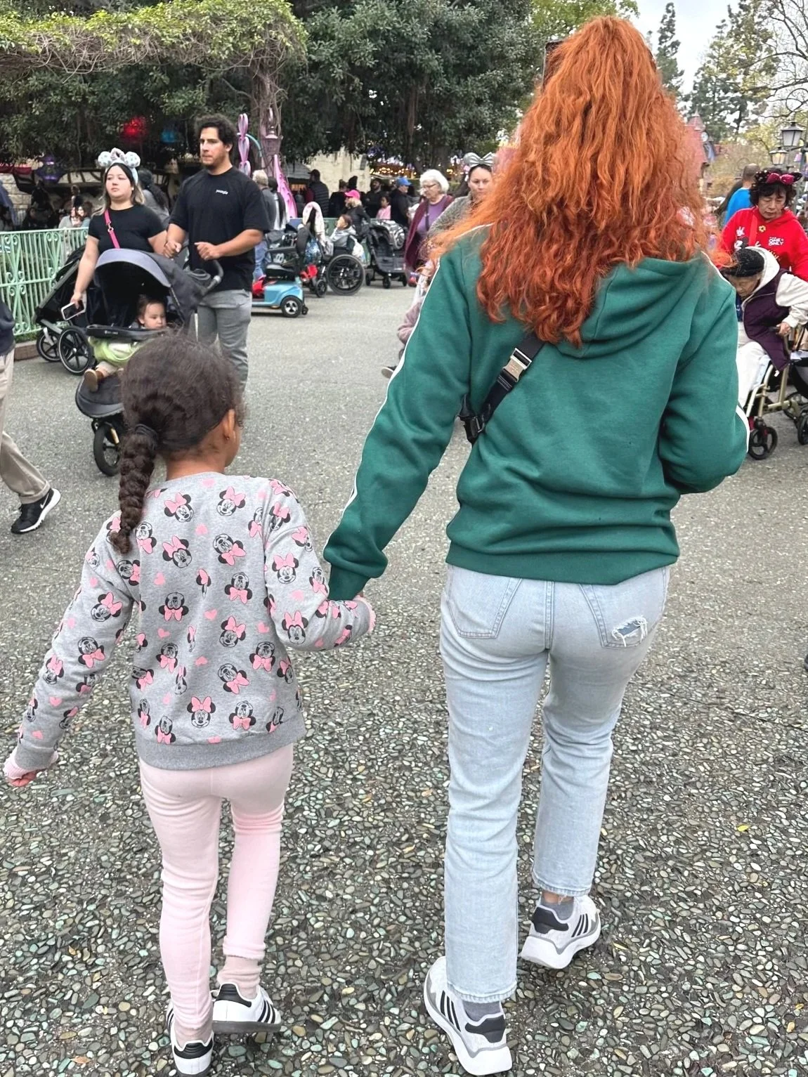 A woman with long red hair holding hands with a young girl at an outdoor amusement park or fair.