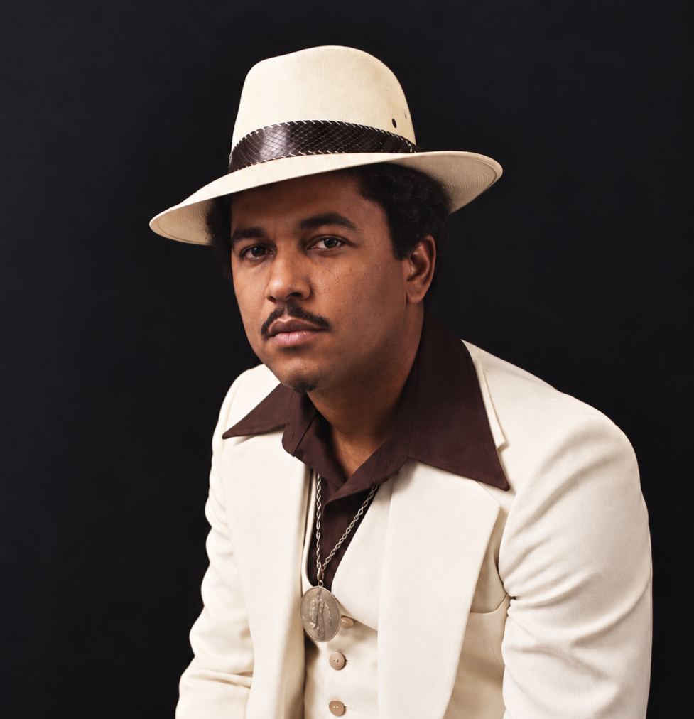 Portrait of Willie Tee wearing a white suit and fedora against a dark background.