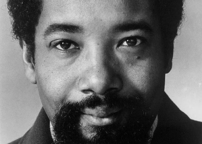 Close-up black-and-white portrait of a man with curly hair, a mustache, and a beard, looking directly at the camera.
