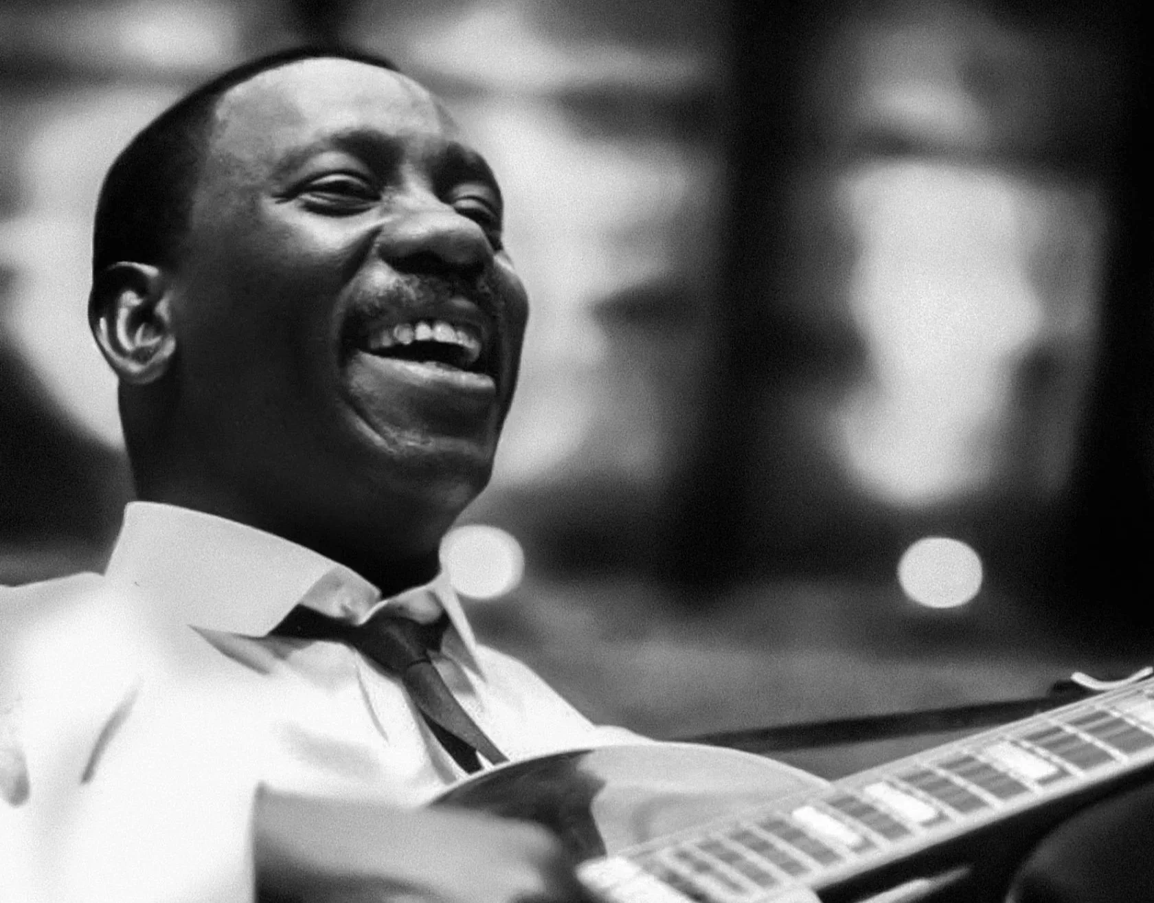 Black-and-white photo of jazz guitarist Wes Montgomery smiling while playing guitar, wearing a white shirt and dark tie.