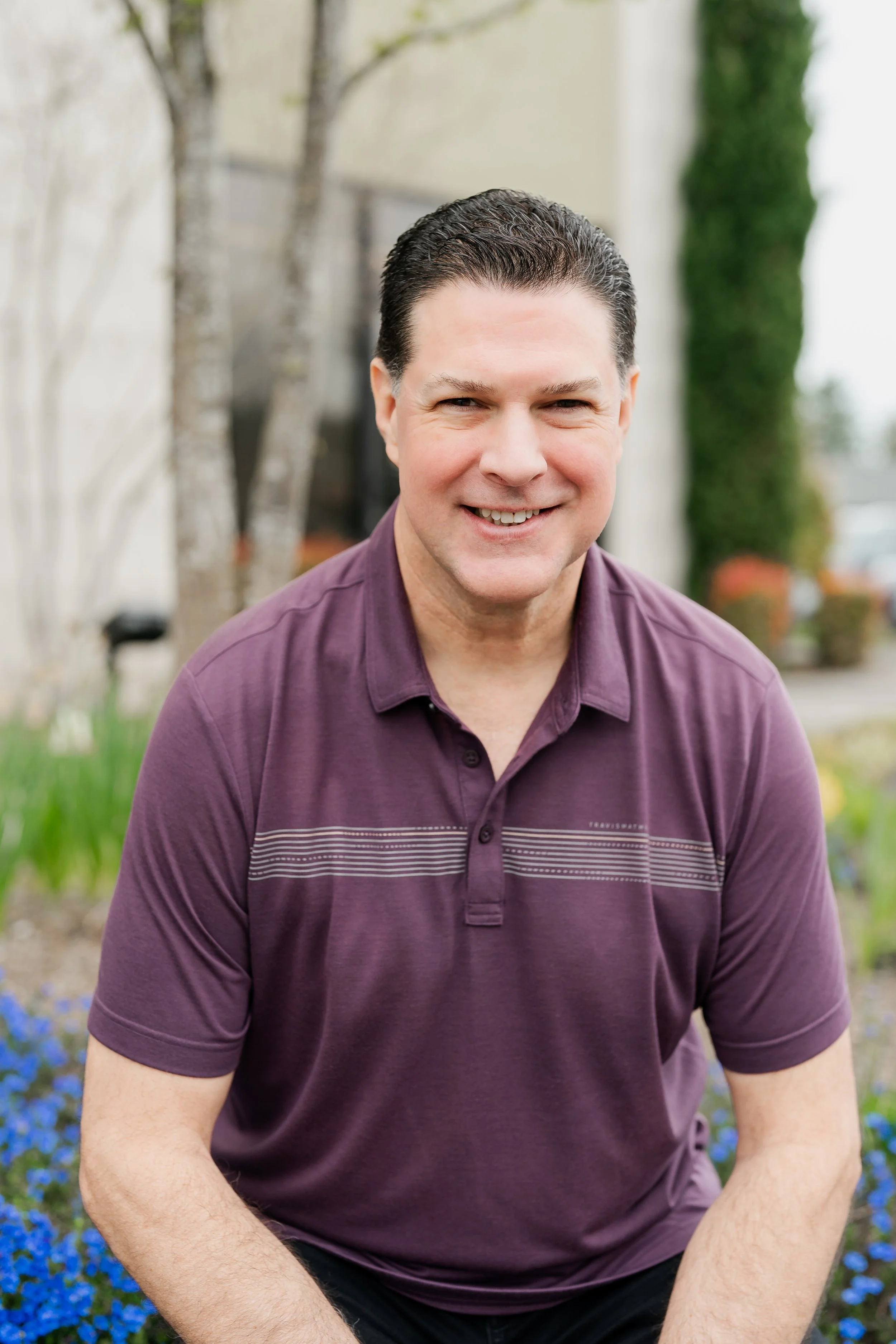 A smiling man with dark hair wearing a purple polo shirt outdoors, with trees and flowers in the background.