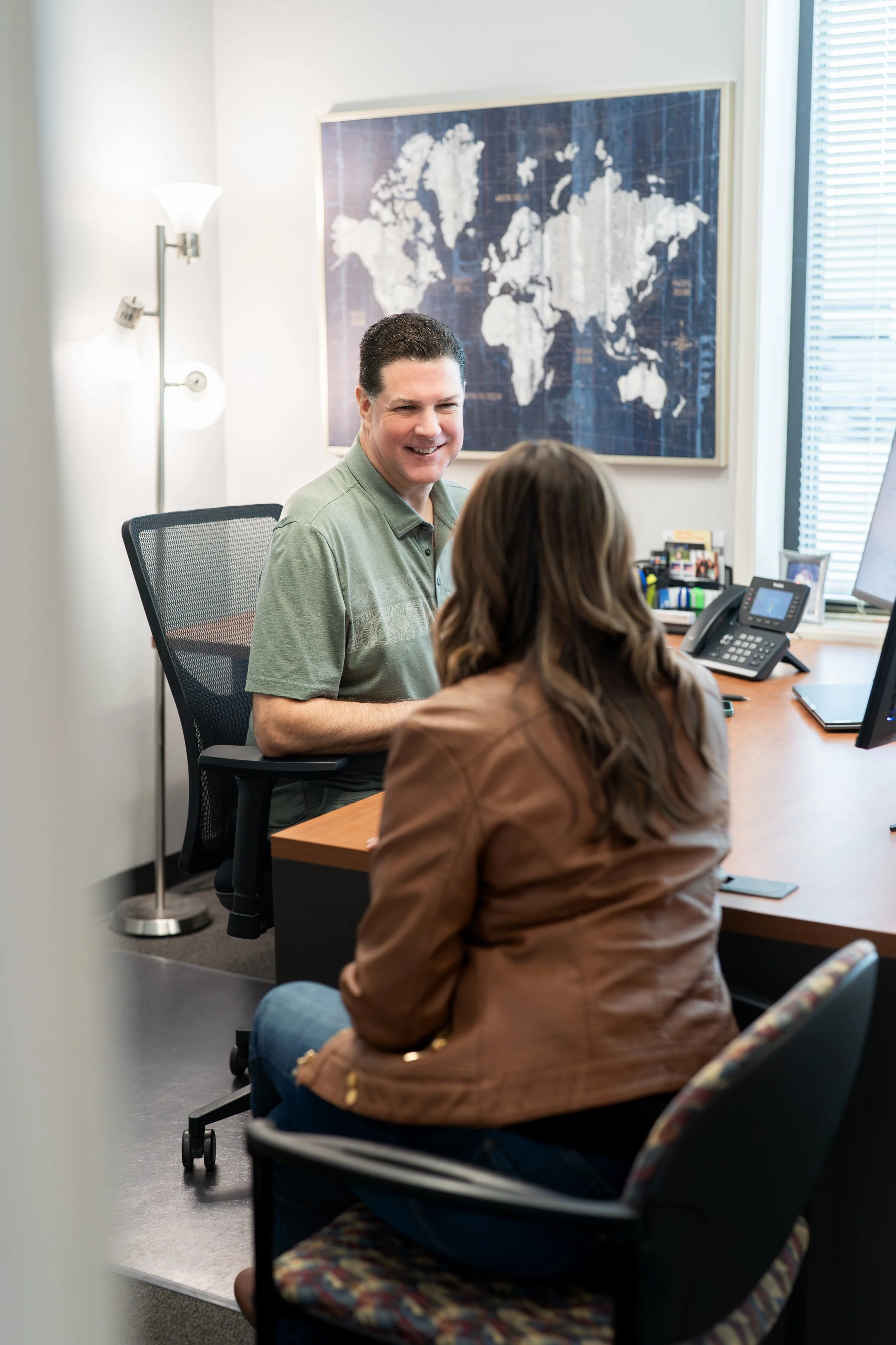 A man smiling while talking to a woman in an office, with a world map on the wall behind them, a lamp, and office supplies on the desk.
