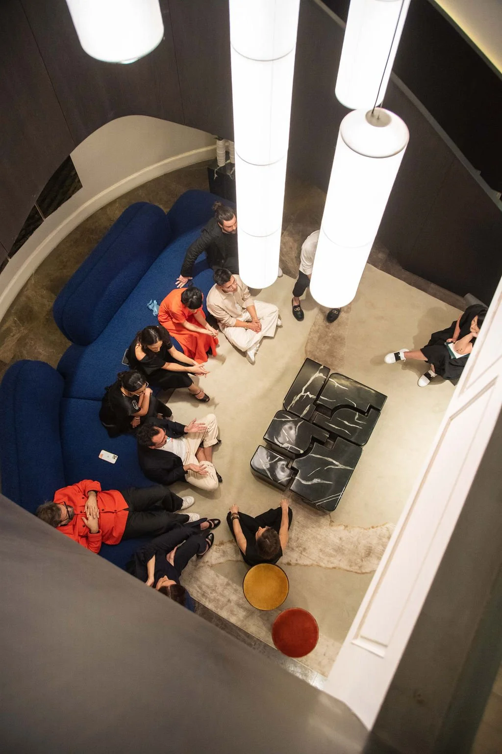 People gathered in a modern, stylish lounge area with blue curved couches and black marble tables, seen from above. White cylindrical pendant lights hang from the ceiling, illuminating the group during a social event or meeting.