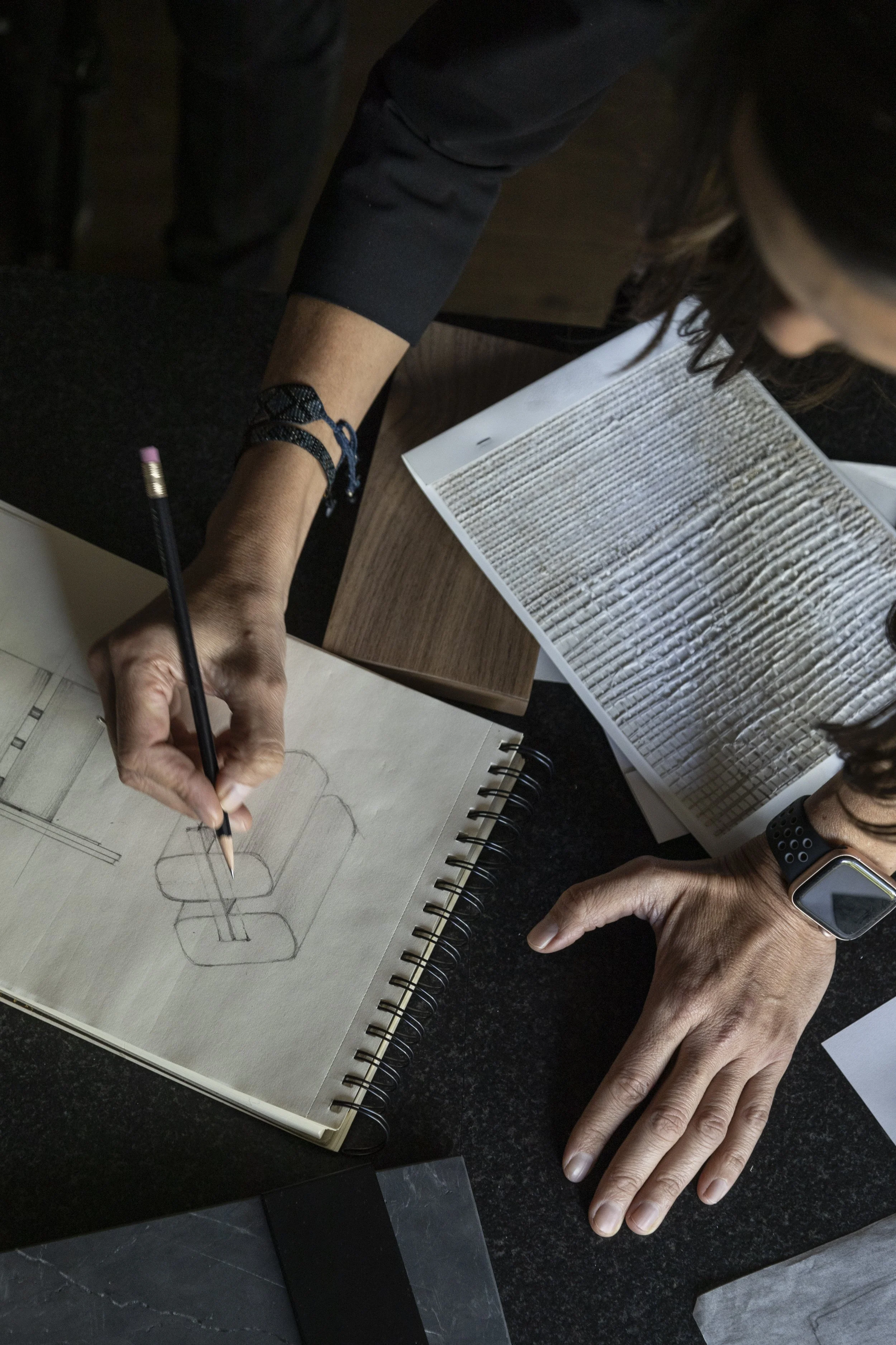People are working on a design sketch of an office chair in a notebook, with one person drawing and another person pointing at the sketch.