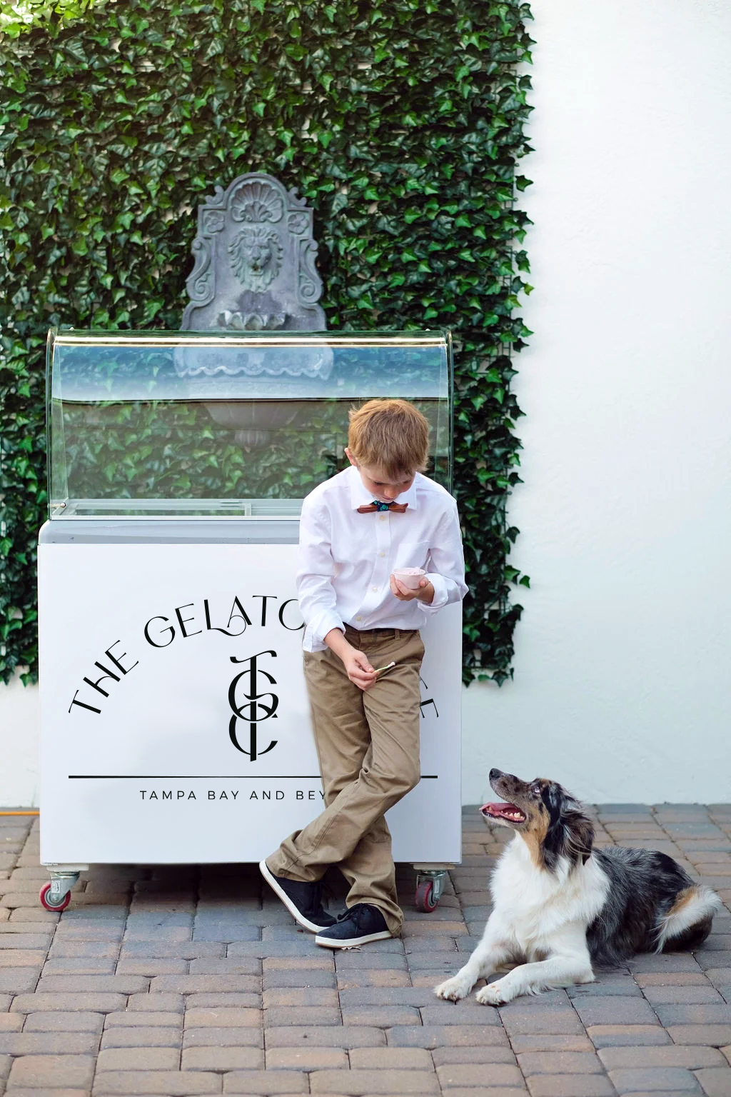 A young boy in a white shirt with a bow tie and khakis stands next to a dog sitting on the brick ground, both looking at an ice cream cone the boy is holding, in front of an ice cream shop with a green ivy-covered wall and a decorative lion fountain in the background.