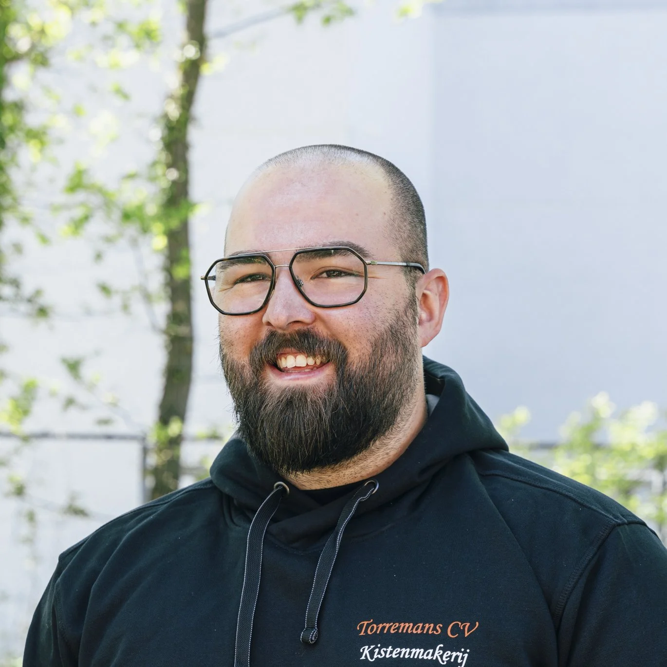 A smiling man with a bald head, glasses, a full beard, and a mustache, wearing a black hoodie with orange and white text, standing outdoors with trees and a building in the background.