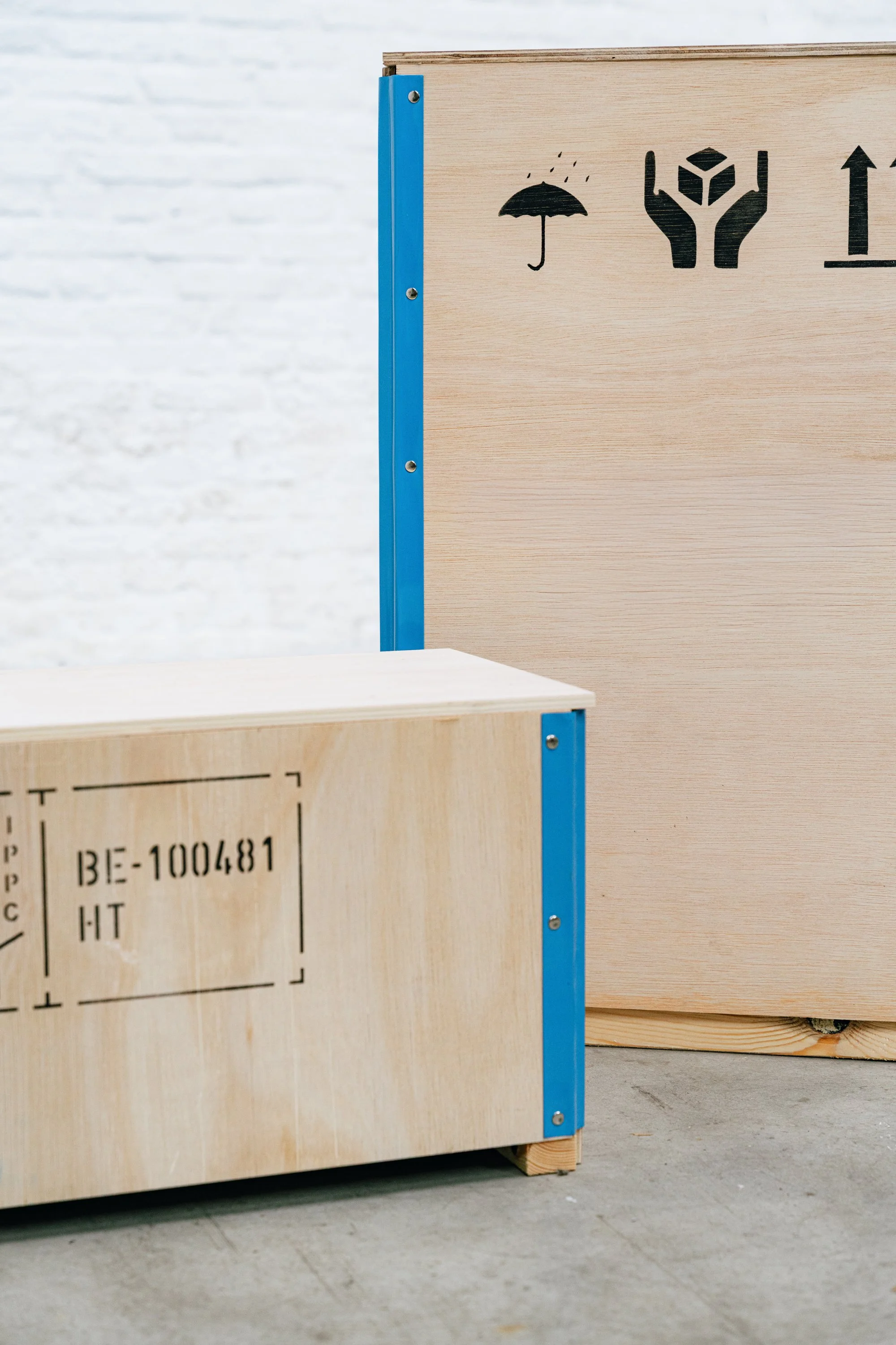 Close-up van houten plooikisten met verzendlabels, metalen hoekbeugels en blauwe metalen steunen, op een betonnen vloer tegen een witte bakstenen muur.