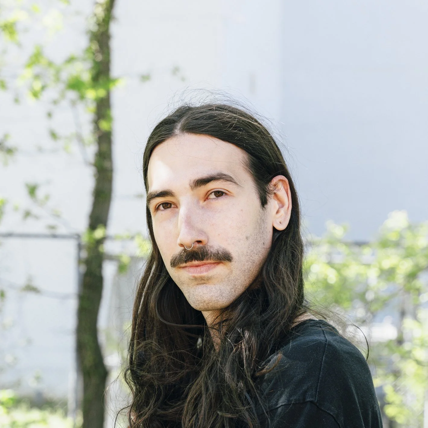 Close-up of a young man with long, dark hair, a mustache, and a septum piercing, outdoors with blurry green trees and blue sky in the background.