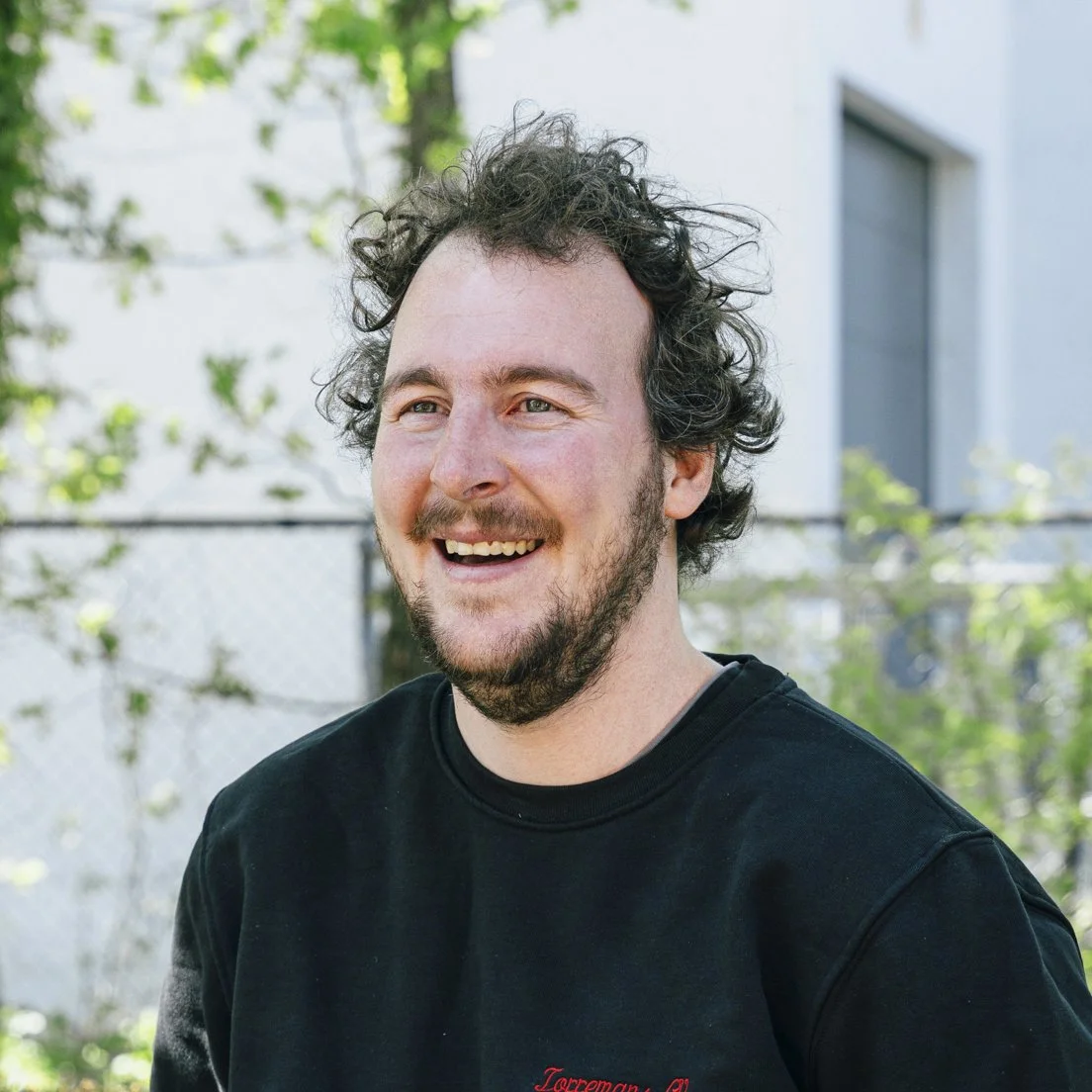 A smiling man with curly dark hair and a beard, wearing a black T-shirt, outdoors with a background of plants, trees, and a building.