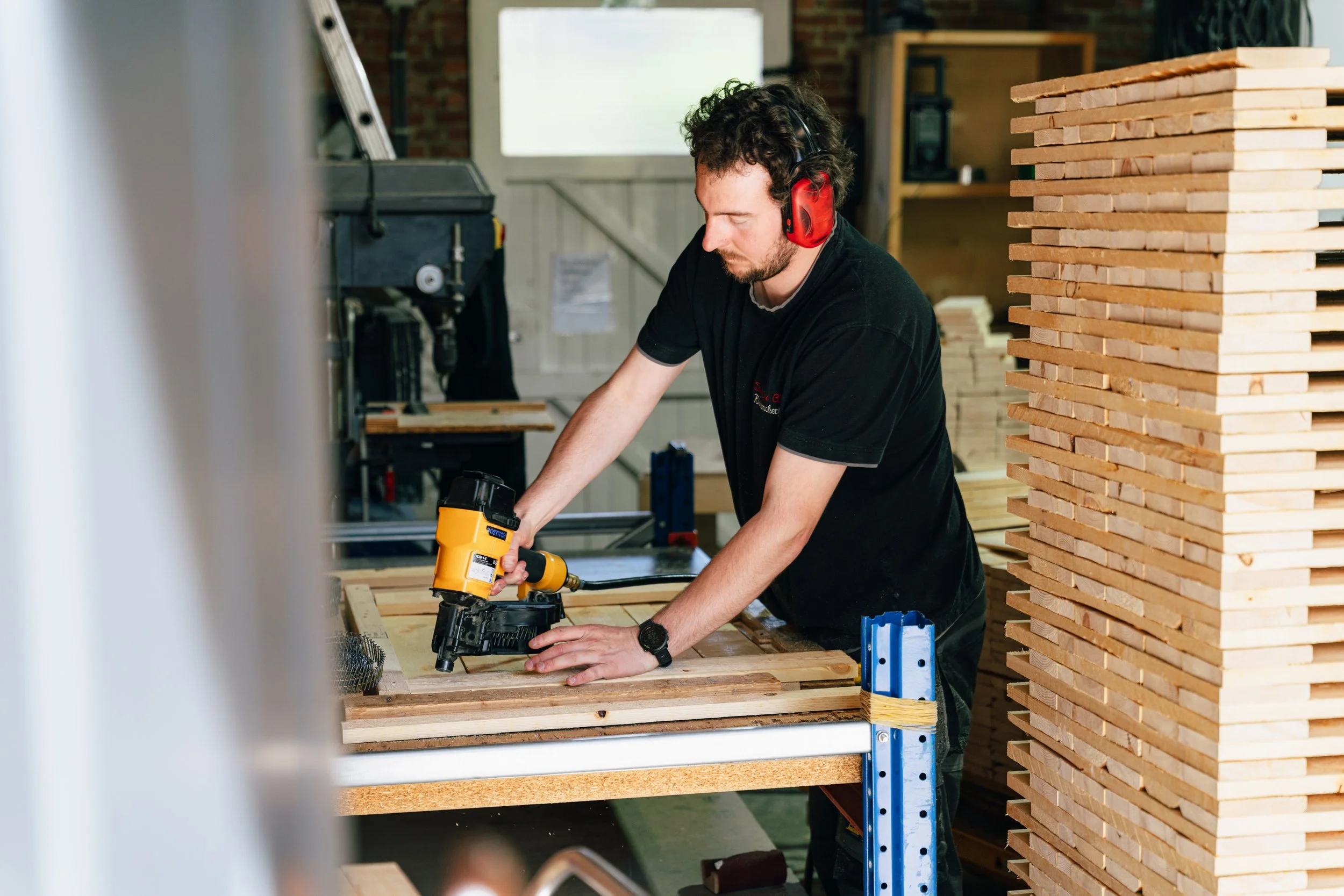 Een werknemer in het gezellig houtbewerkingsate in Herentals met een zwart T-shirt en gebruikt een spijkerpistool om een houten onderdeel samen te stel…Een man met rode gehoorbescherming en een zwart T-shirt werkt i