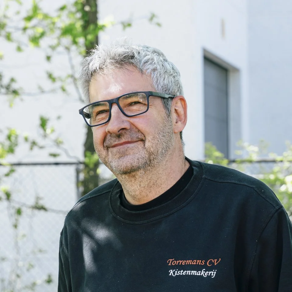 Oprichter Manno Torremans met grijs haar en baard, een bril en een zwarte sweater met de tekst 'Torremans CV Kistenmakerij', glimlachend buiten voor een wit gebouw met groen