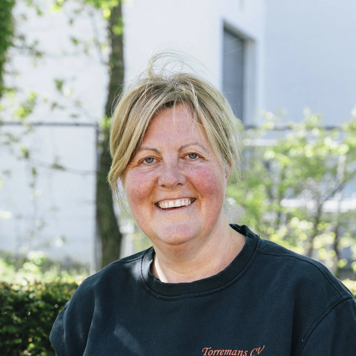 Werkneemster glimlachende met kort blond haar, gekleed in een zwarte sweater met rode tekst "Torremans CV", staand buiten met bomen en een gebouw op de achtergrond.