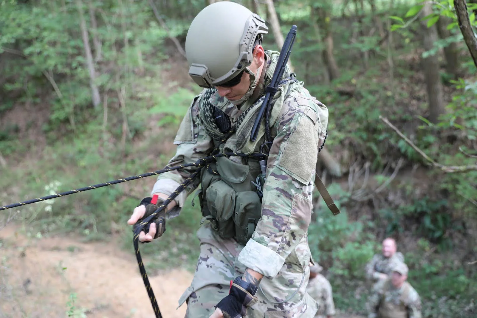 A soldier in camouflage uniform, helmet, sunglasses, and tactical gear, pulling a rope during a training exercise in a wooded area, with other soldiers visible in the background.