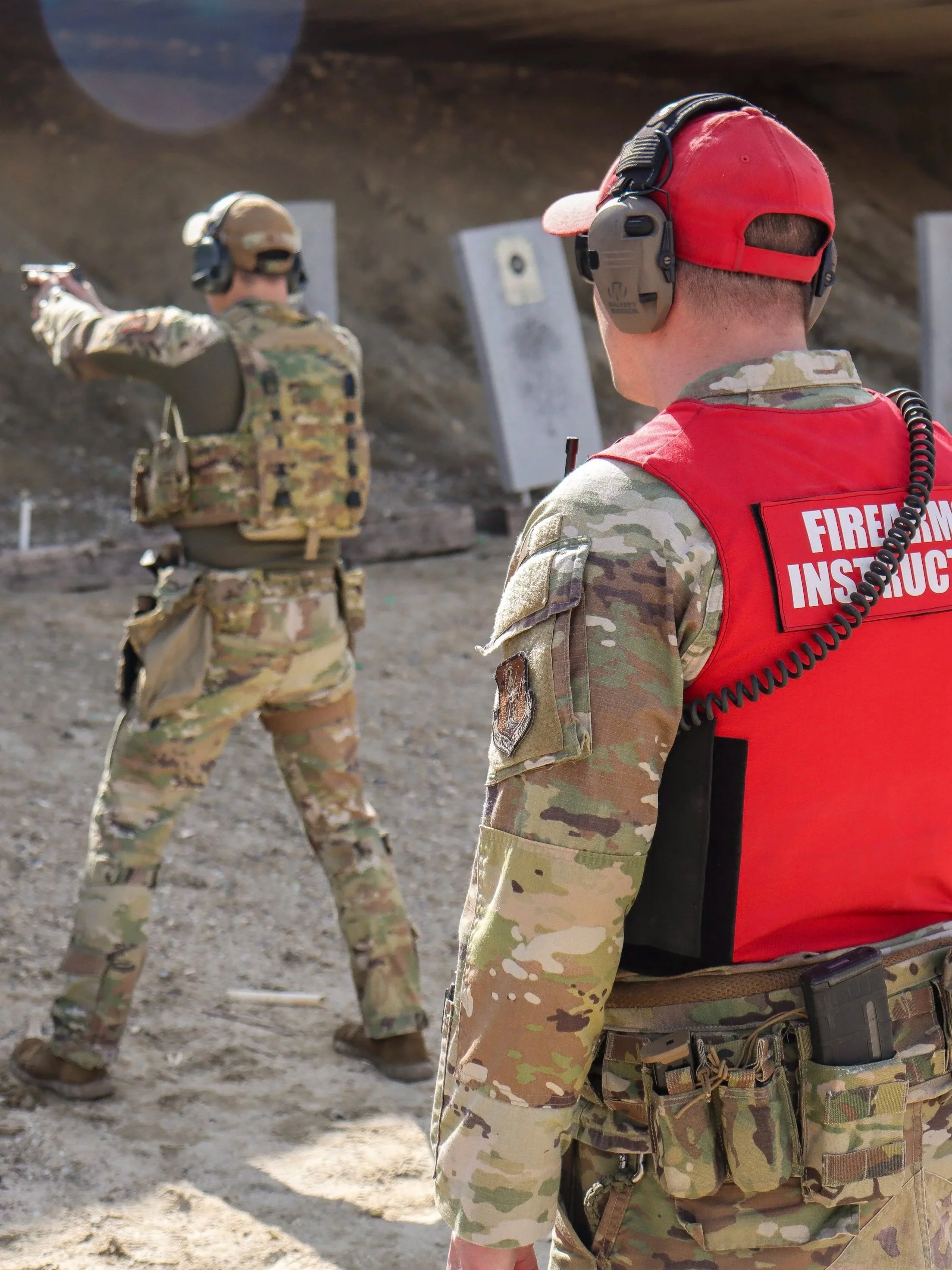 A firearm instructor in camouflage gear watches a person firing at a shooting range outdoors.